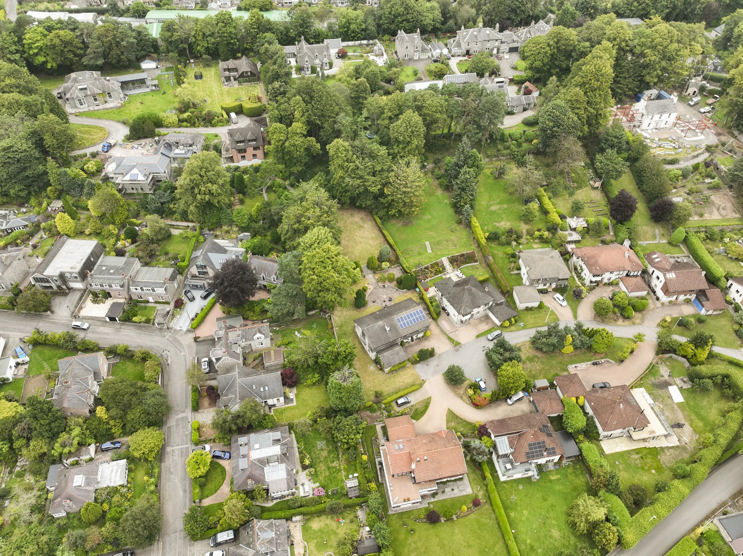 An aerial view of a residential neighborhood with multiple houses, trees, gardens, and streets visible. Some houses have solar panels on their roofs.