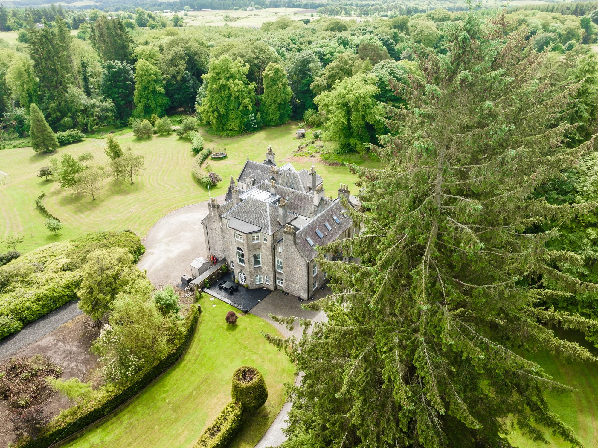Aerial view of a large historic stone mansion surrounded by lush green trees and manicured lawn, with open fields in the background.