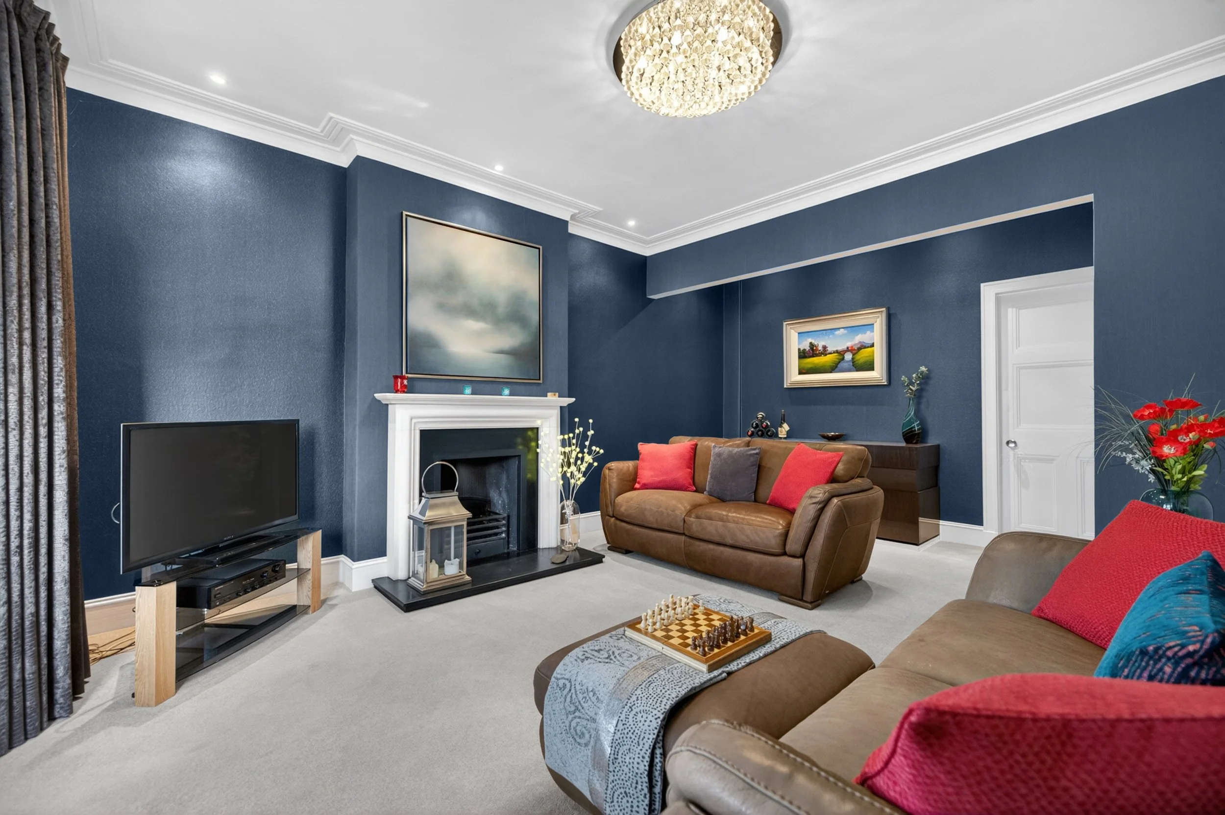 A living room with dark blue walls, a white fireplace, a flat-screen TV on a wooden stand, a leather sofa with multicolored pillows, a coffee table with a chessboard, a decorative plant, and framed landscape paintings on the wall. There is a chandeli