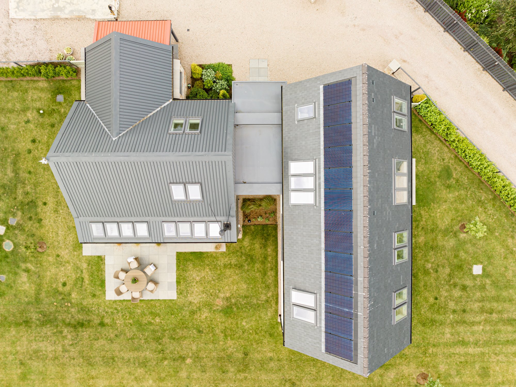An aerial view of a modern house with gray roof, four skylights, solar panels, and surrounding gardens and patio furniture.