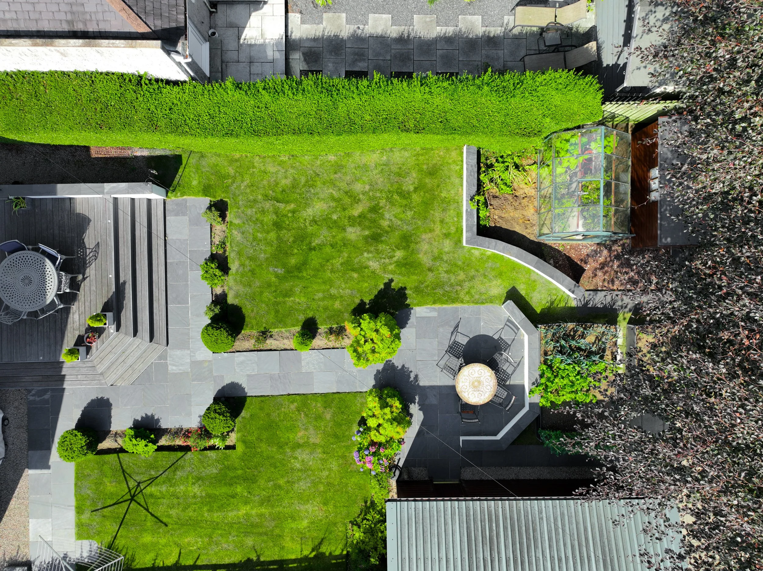 A backyard with a green hedge, a small greenhouse, patio furniture, and a grassy lawn.