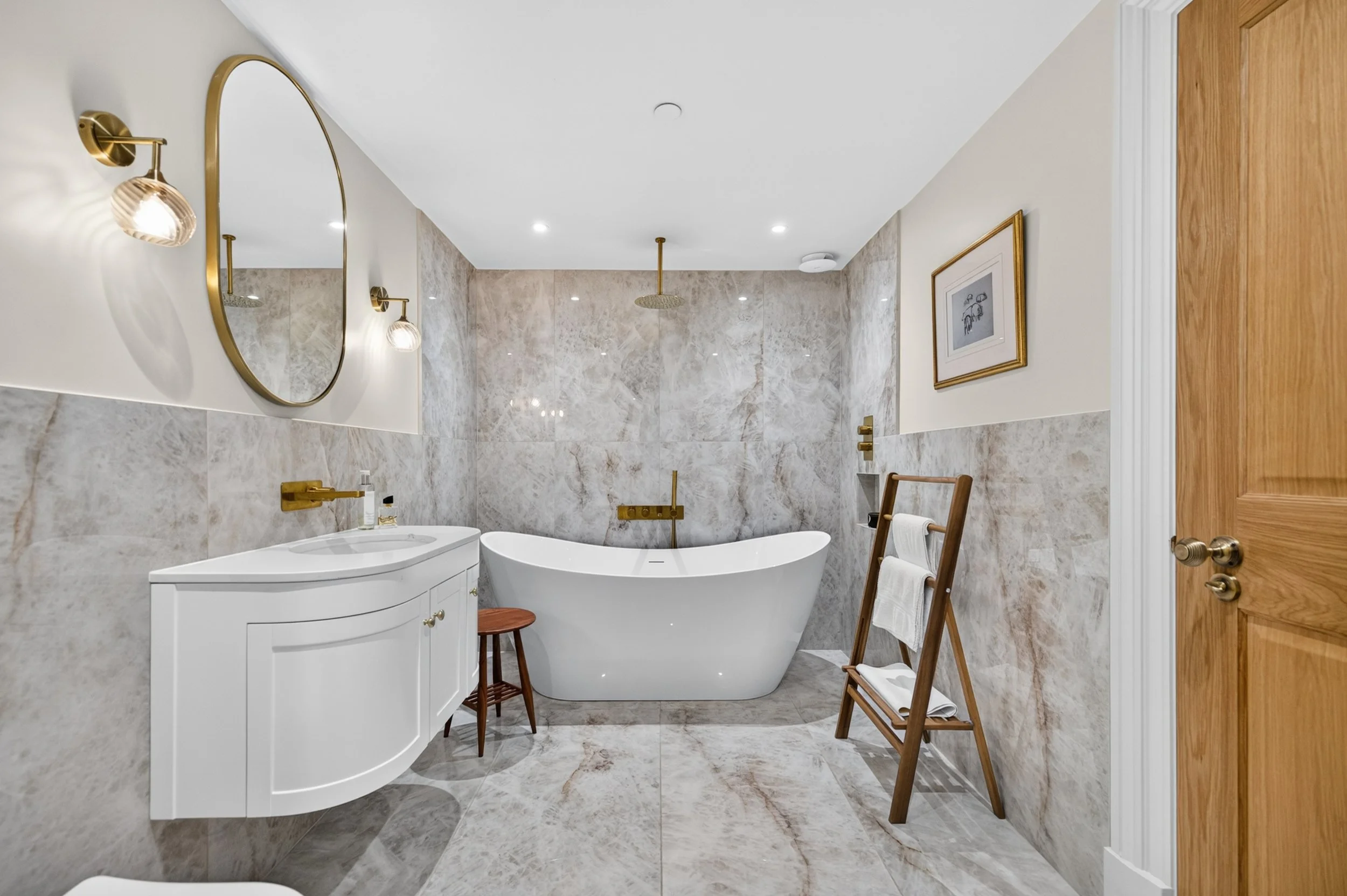 A modern bathroom with a freestanding bathtub, wall-mounted gold fixtures, a white vanity with cabinet storage, a round mirror, wall sconces, marble wall and floor tiles, a framed picture, a wooden door, and a ladder towel rack with white towels.