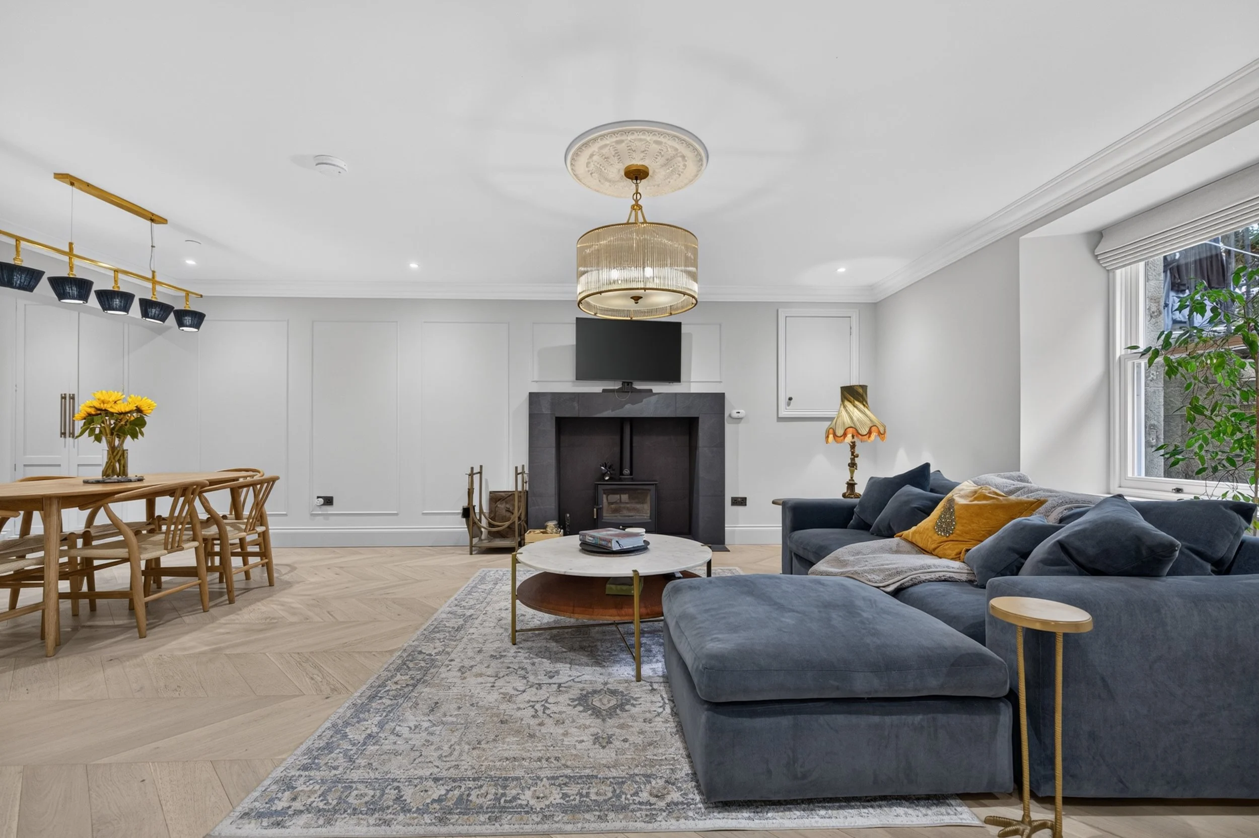 Living room with a blue sofa, a white round coffee table, and a vintage lamp. There is a fireplace with a TV above it, a dining table with wooden chairs, a chandelier, and a window with blinds and a potted plant.