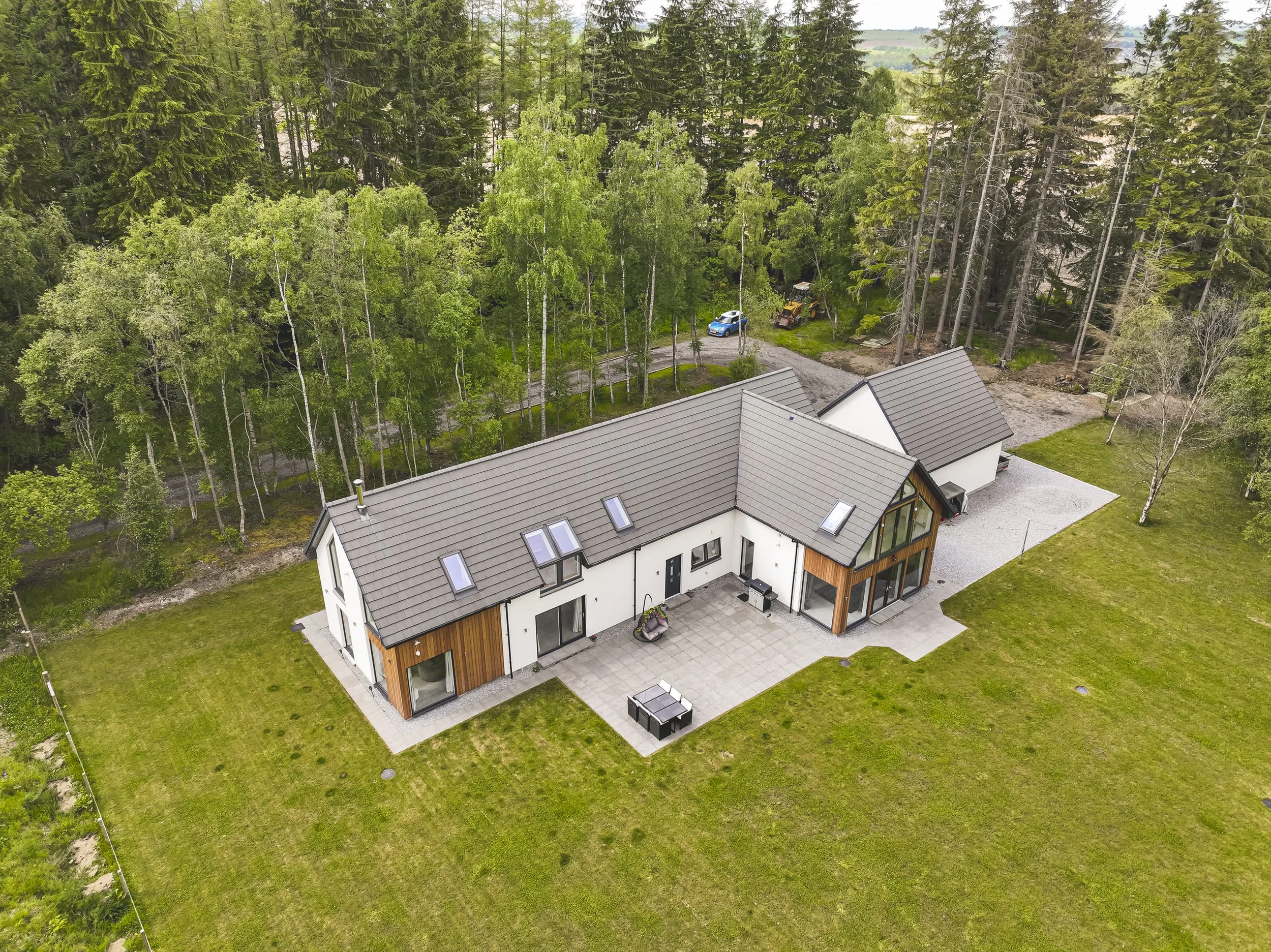 An aerial view of a modern house with a gray roof, large windows, and a backyard patio, surrounded by green grass and tall trees.