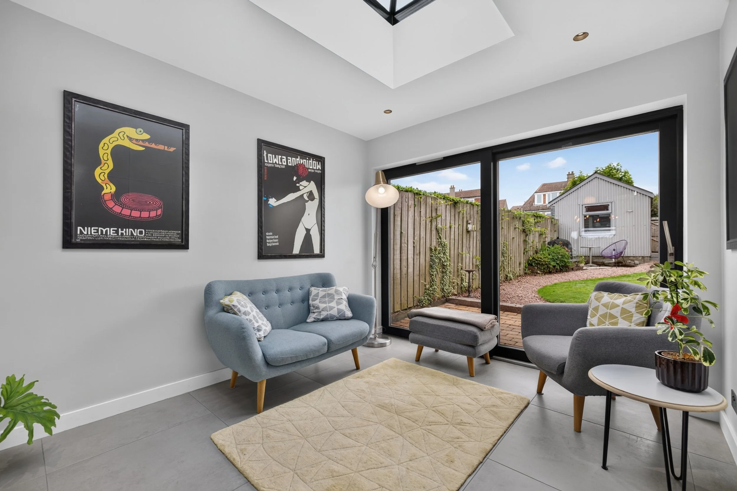 Living room with large sliding glass doors leading to a backyard, light gray walls, framed artwork on the wall, a gray armchair with white patterned pillows, a small side table with a potted plant, and a beige rug on the gray tile floor. Outside view shows a wooden fence, garden, and neighboring house.