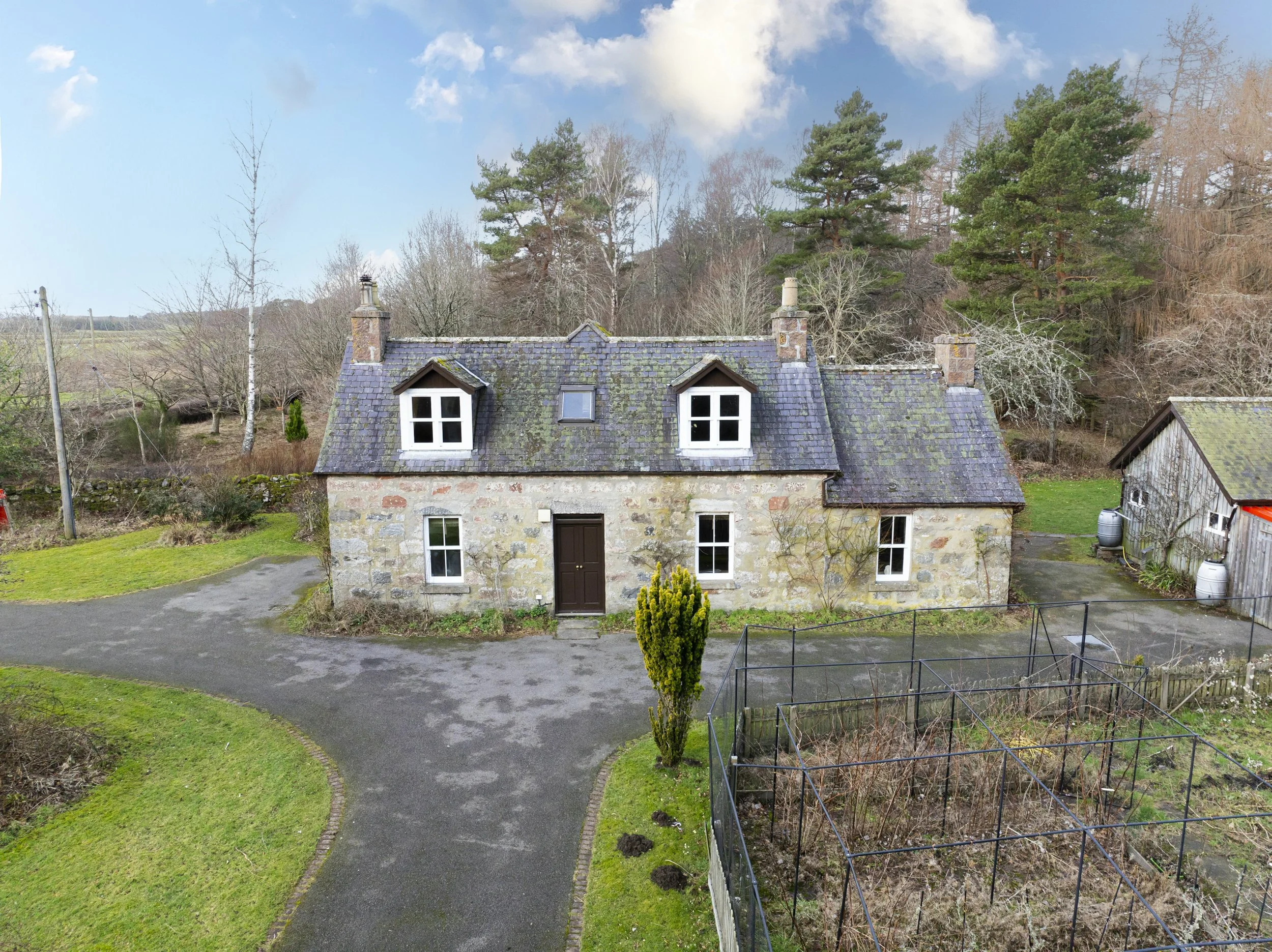 A stone house with a slate roof, white framed windows, and two brick chimneys, surrounded by a garden with a curved pathway, leafless trees, and greenery, with a backdrop of tall pine trees and a partly cloudy sky.