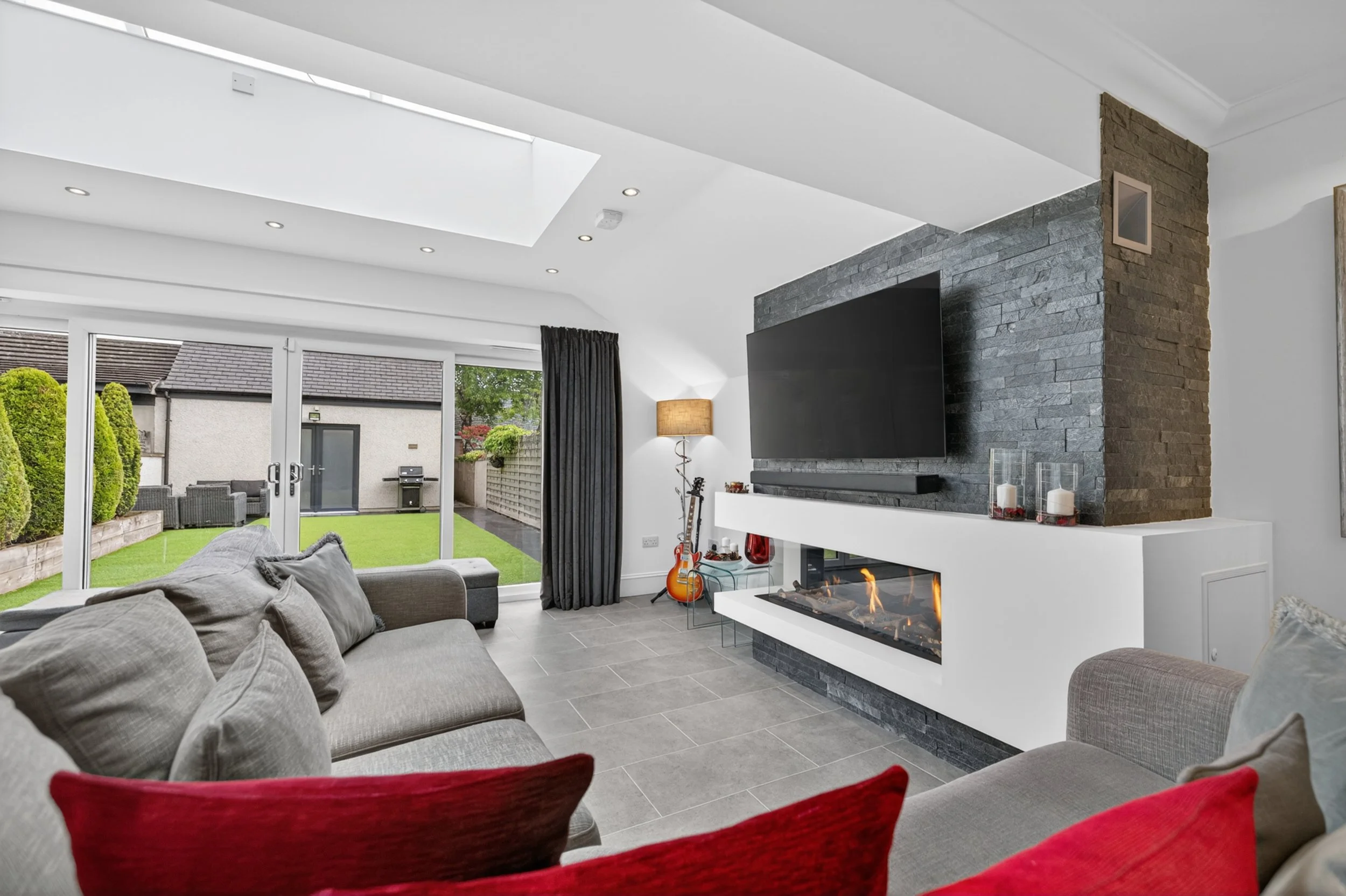 Modern living room with gray and red seating, a fireplace, a wall-mounted TV, and large glass doors opening to a backyard with a lawn and outdoor furniture.
