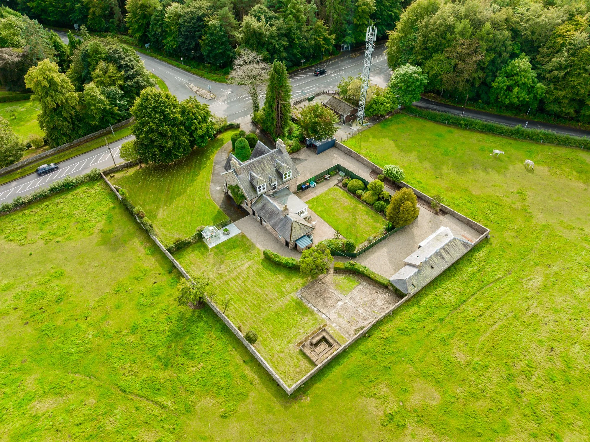 An aerial view of a rural scene with a large house and a fenced yard, green fields, and a forest in the background. There are a few horses grazing on the grass field.