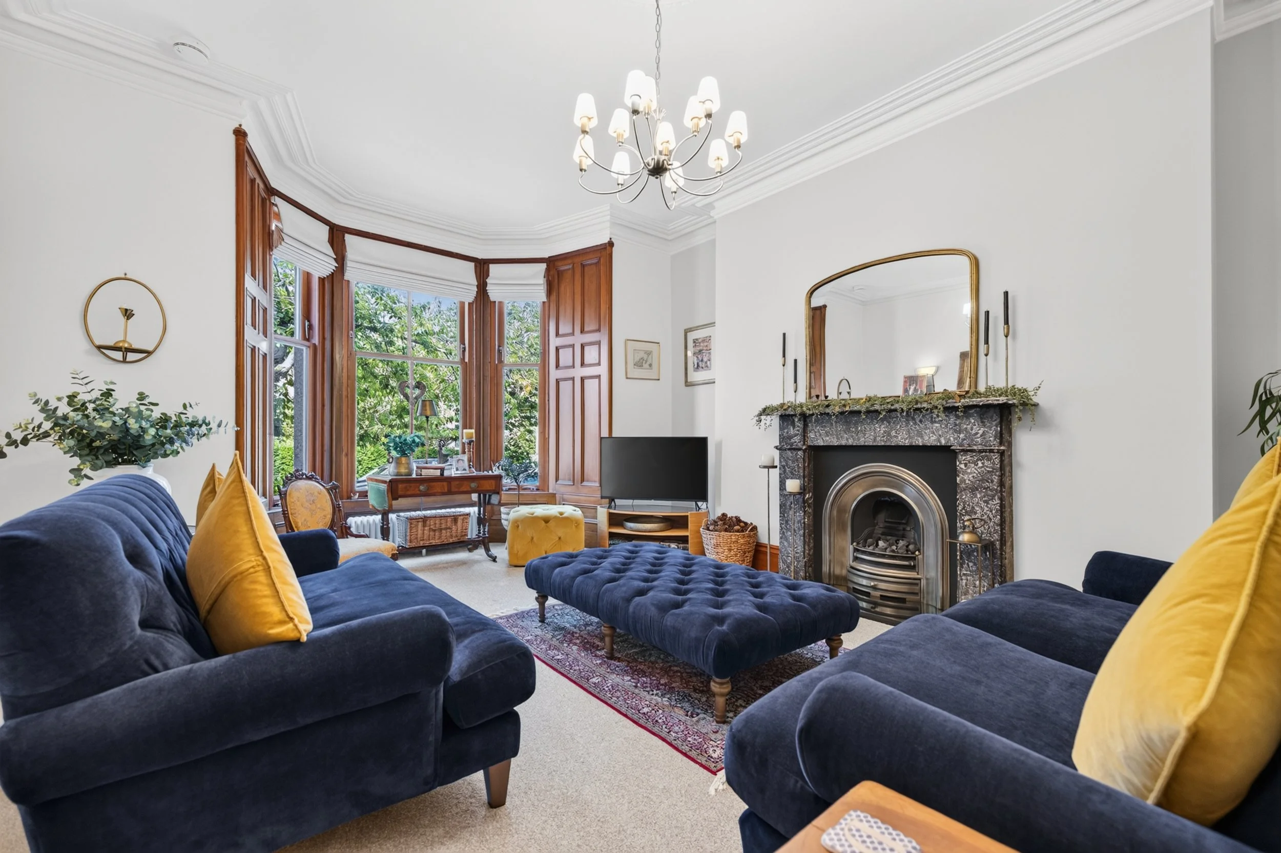A living room with a velvet navy blue sofa and matching ottoman, mustard yellow pillows, a fireplace with a mirror, a television, large bay windows with wooden frames, and various decorative items.