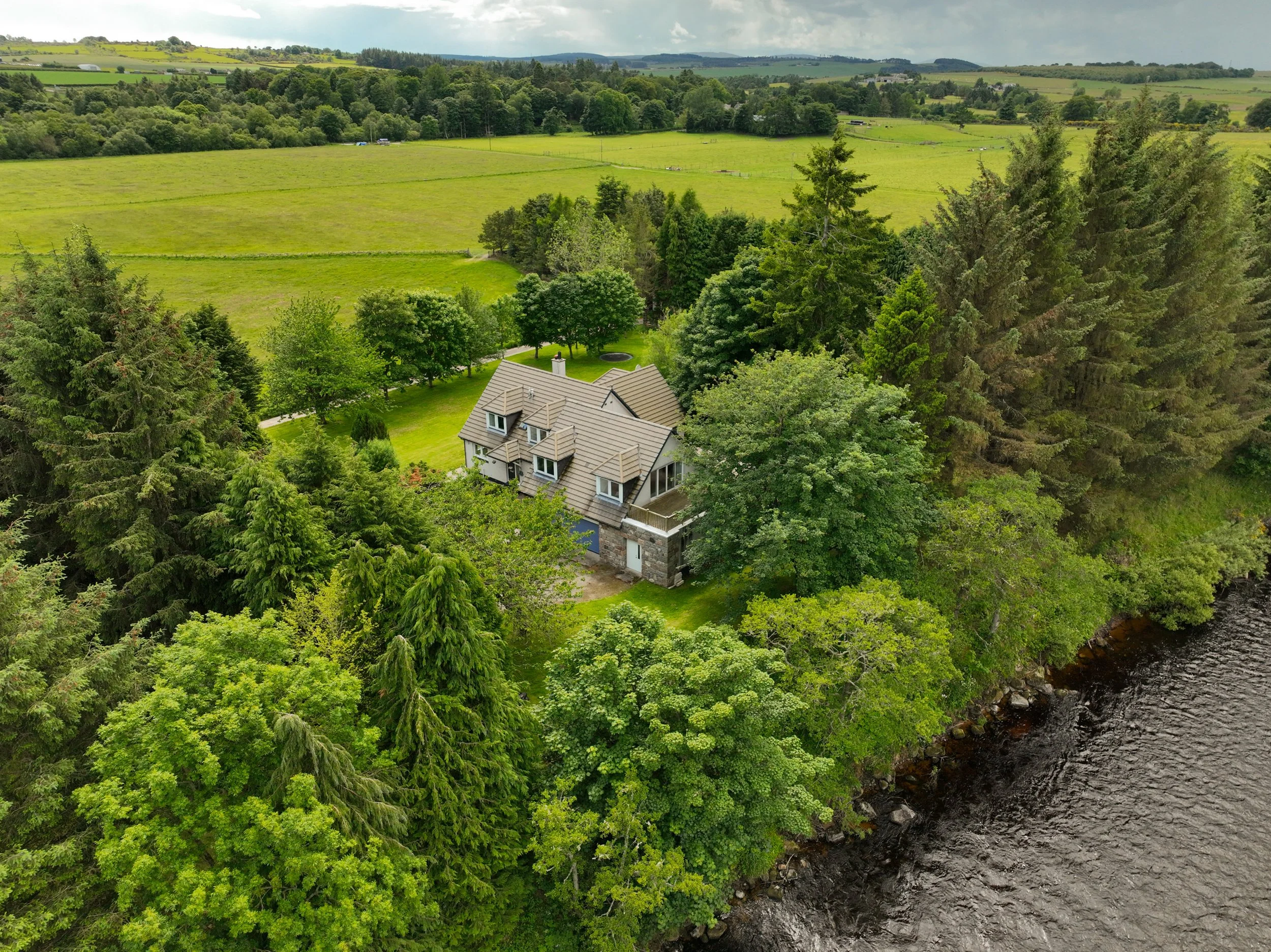 Aerial view of a house surrounded by lush green trees, open fields, and a river in a countryside landscape under a partly cloudy sky.