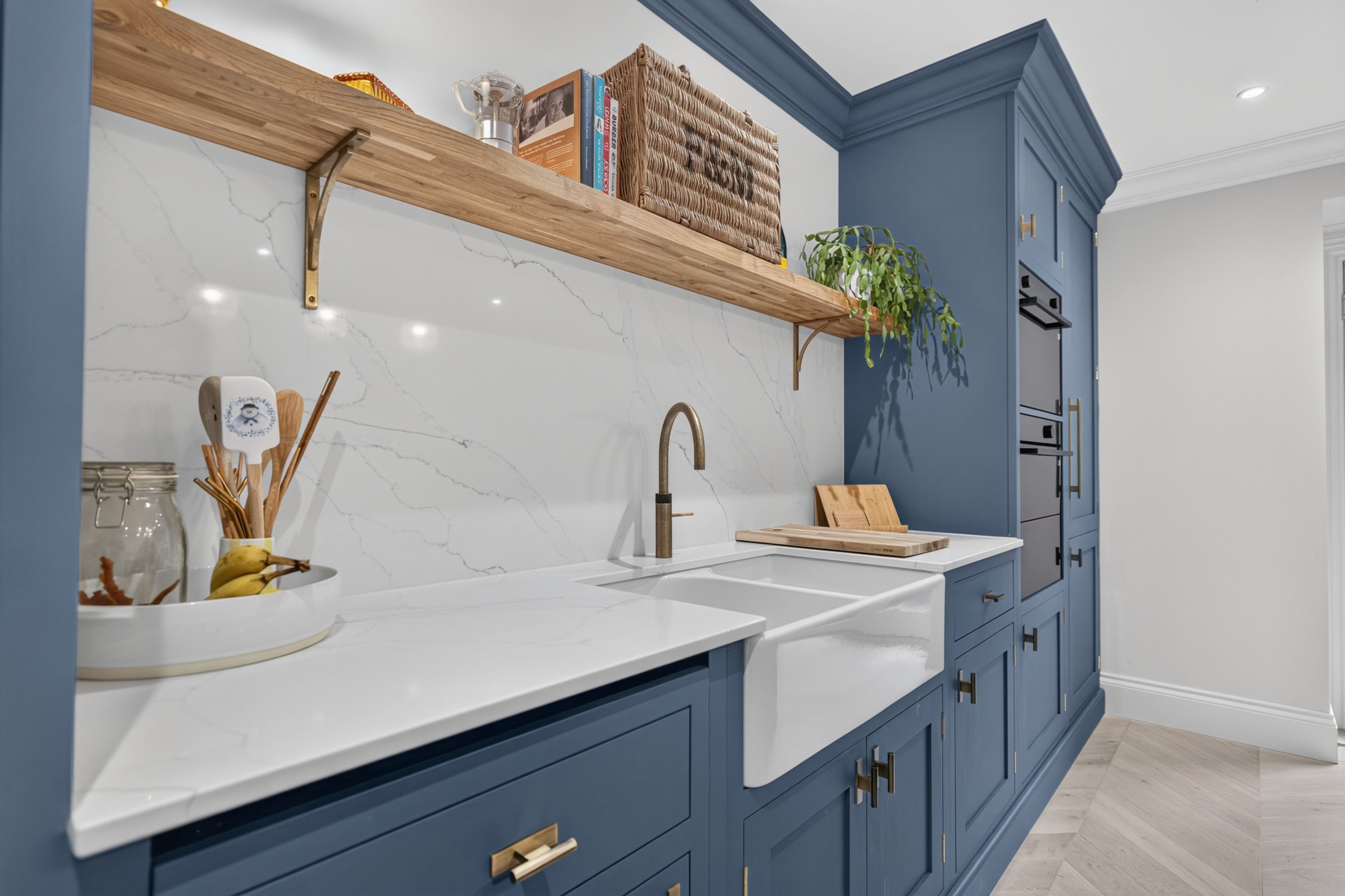 Kitchen with blue cabinets, white marble countertop, and open shelving. Items include a plant, books, a basket, and kitchen utensils on the counter, with an undermount sink and brass hardware.
