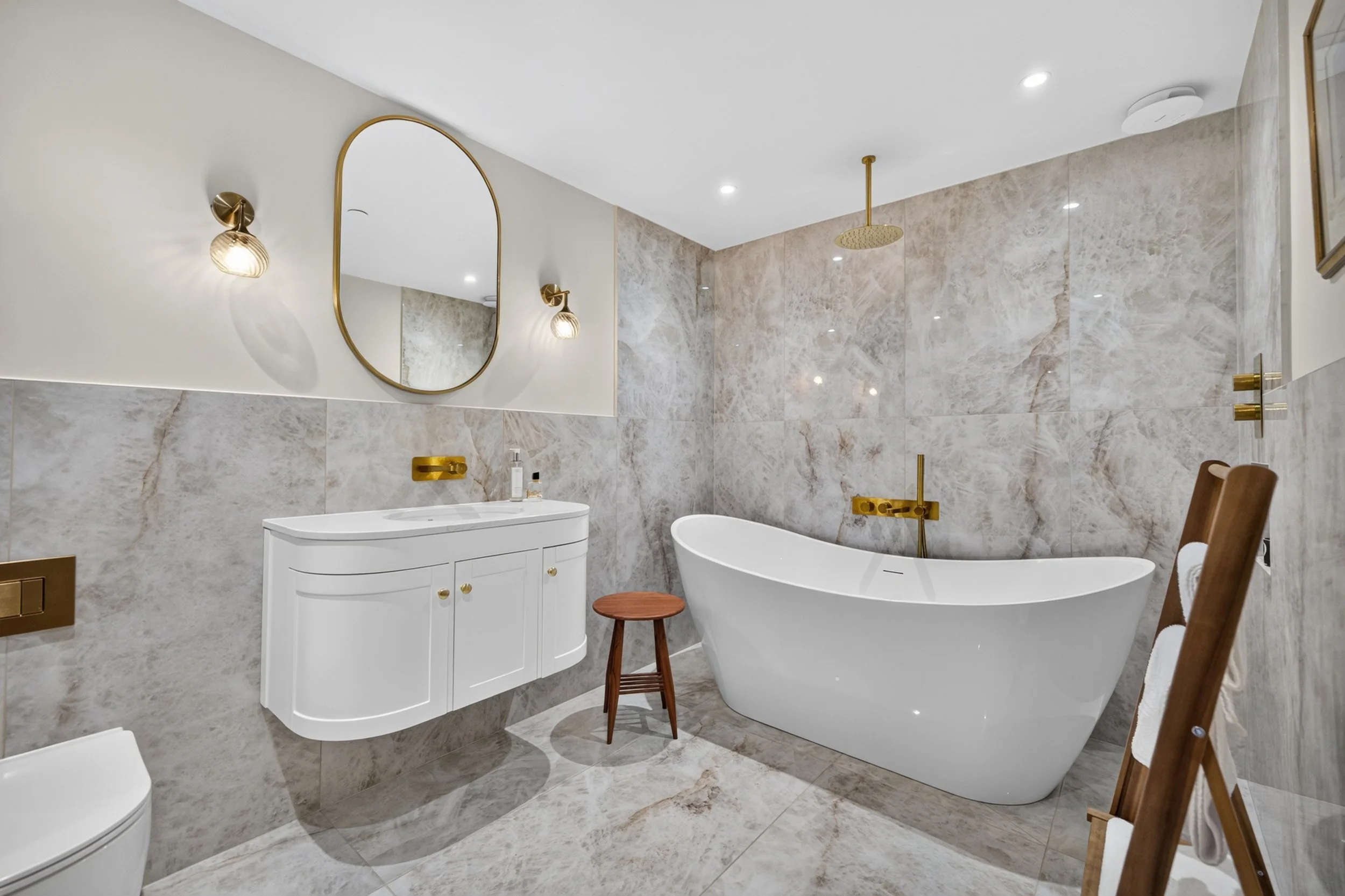 Modern bathroom with marble tiles, white freestanding bathtub, wooden stool, white curved vanity with gold fixtures, oval mirror, wall-mounted lights, shower head, and wooden chairs.