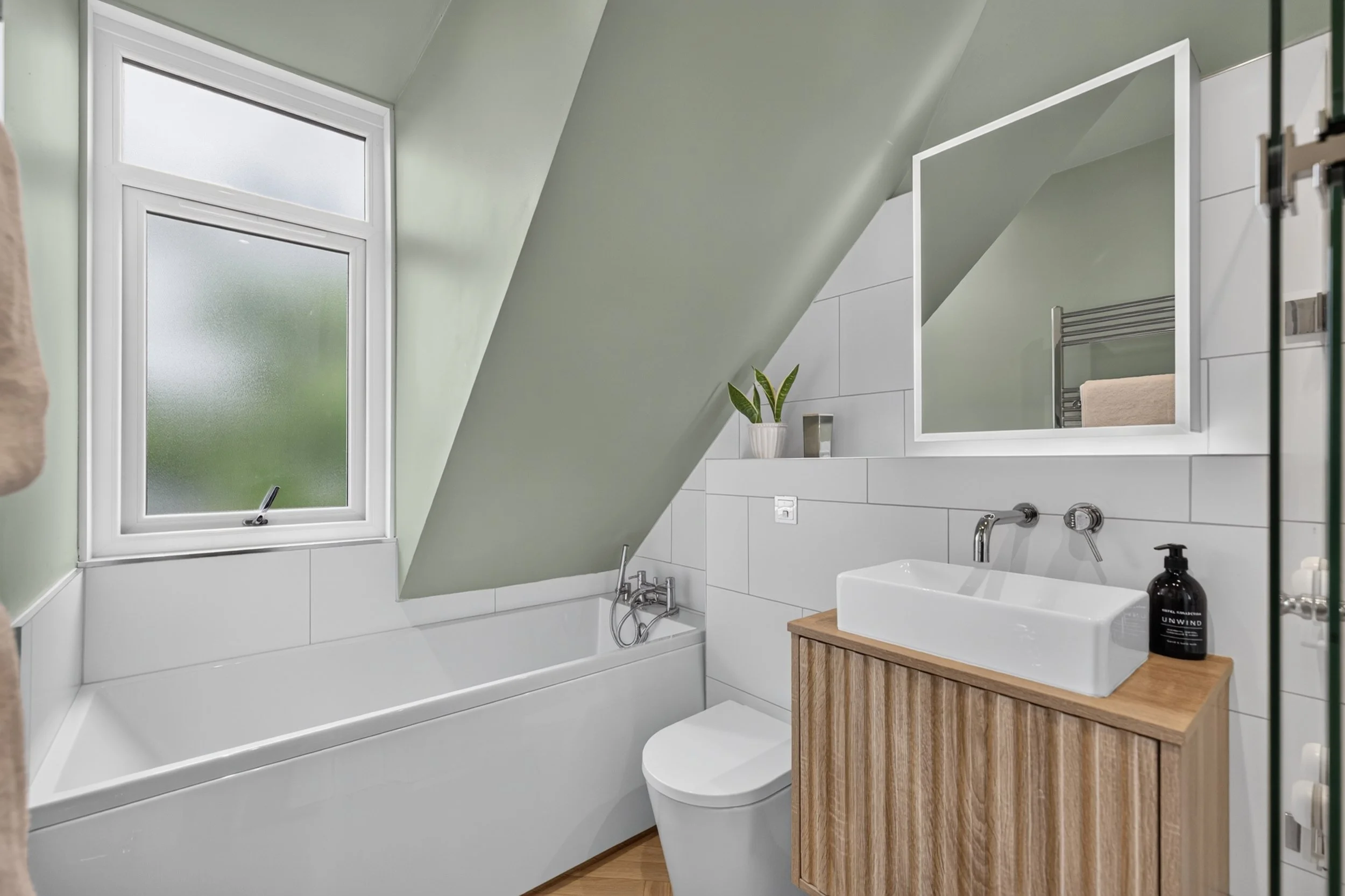 A bathroom with a large frosted window, white bathtub, modern sink with a wooden cabinet, mirror, and minimal decor including a potted plant and soap dispenser.