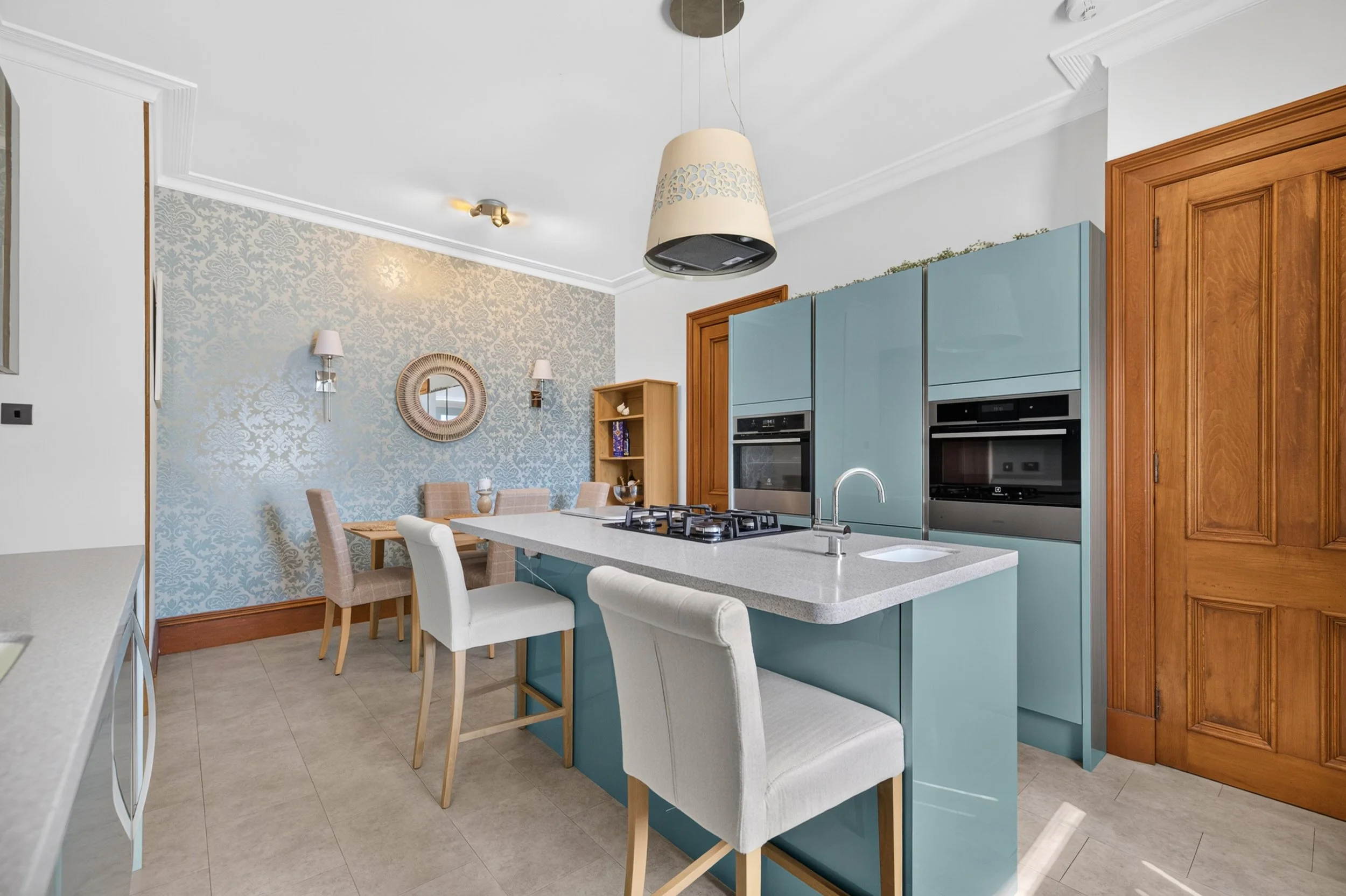 Modern kitchen with light blue cabinets, a white kitchen island, beige chairs, wooden door, patterned wallpaper, round mirror, wall sconces, and a ceiling light fixture.