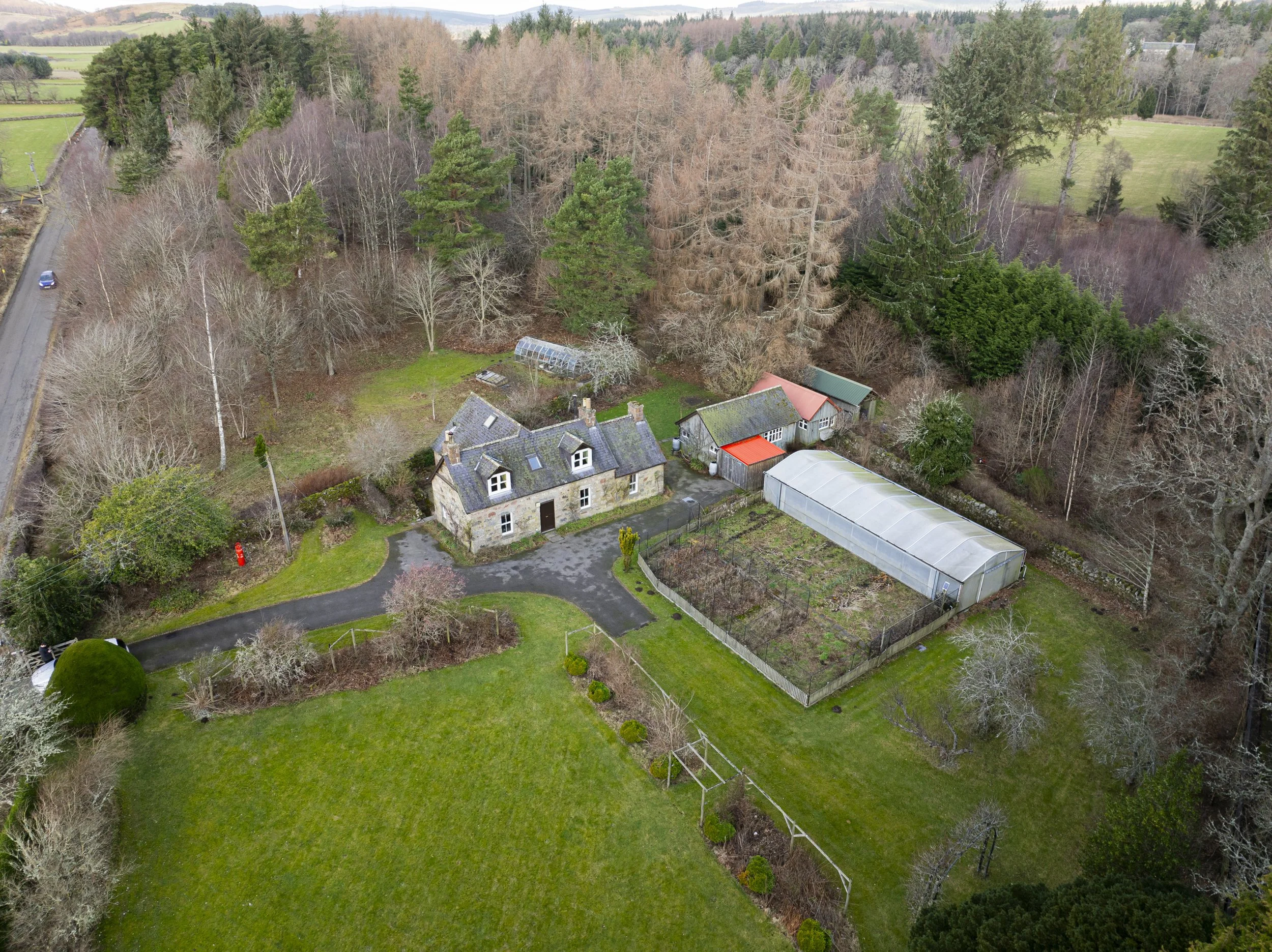 An aerial view of a countryside property with a stone house, green lawns, a greenhouse, and surrounding trees, with a rural road on the left and fields in the distance.