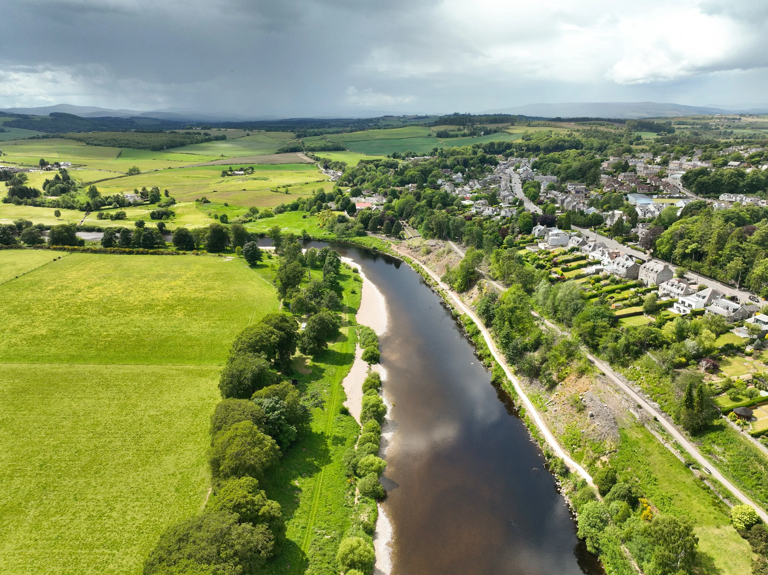 Aerial view of a lush green landscape with a river, a riverbank, a small town, and open fields under a partly cloudy sky.