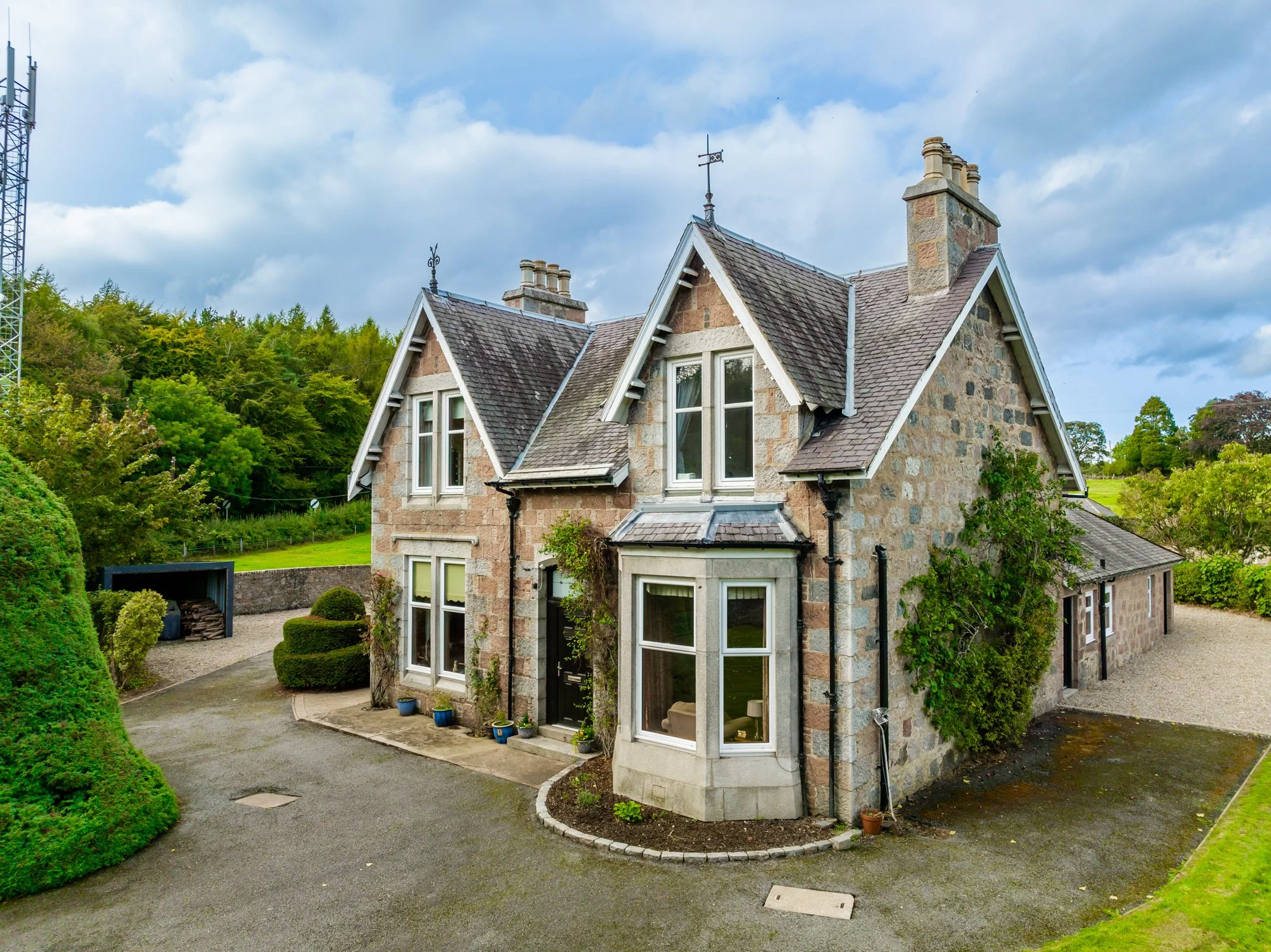 A stone house with a sloped roof, multiple chimneys, and multiple windows, surrounded by greenery and a driveway.