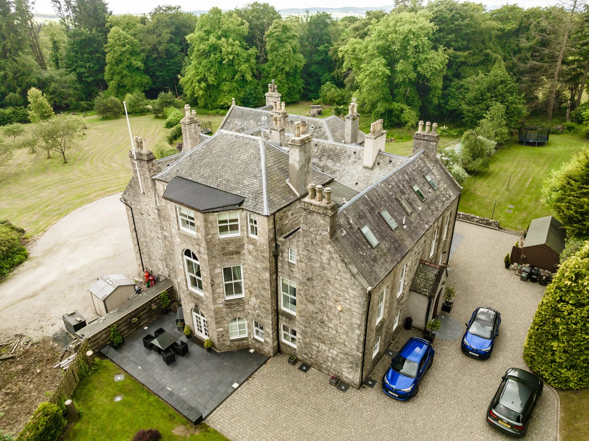 A large stone mansion with multiple chimneys and a slate roof. The house has white framed windows, a patio area with outdoor furniture, surrounded by a green lawn, trees, and a driveway with parked cars.