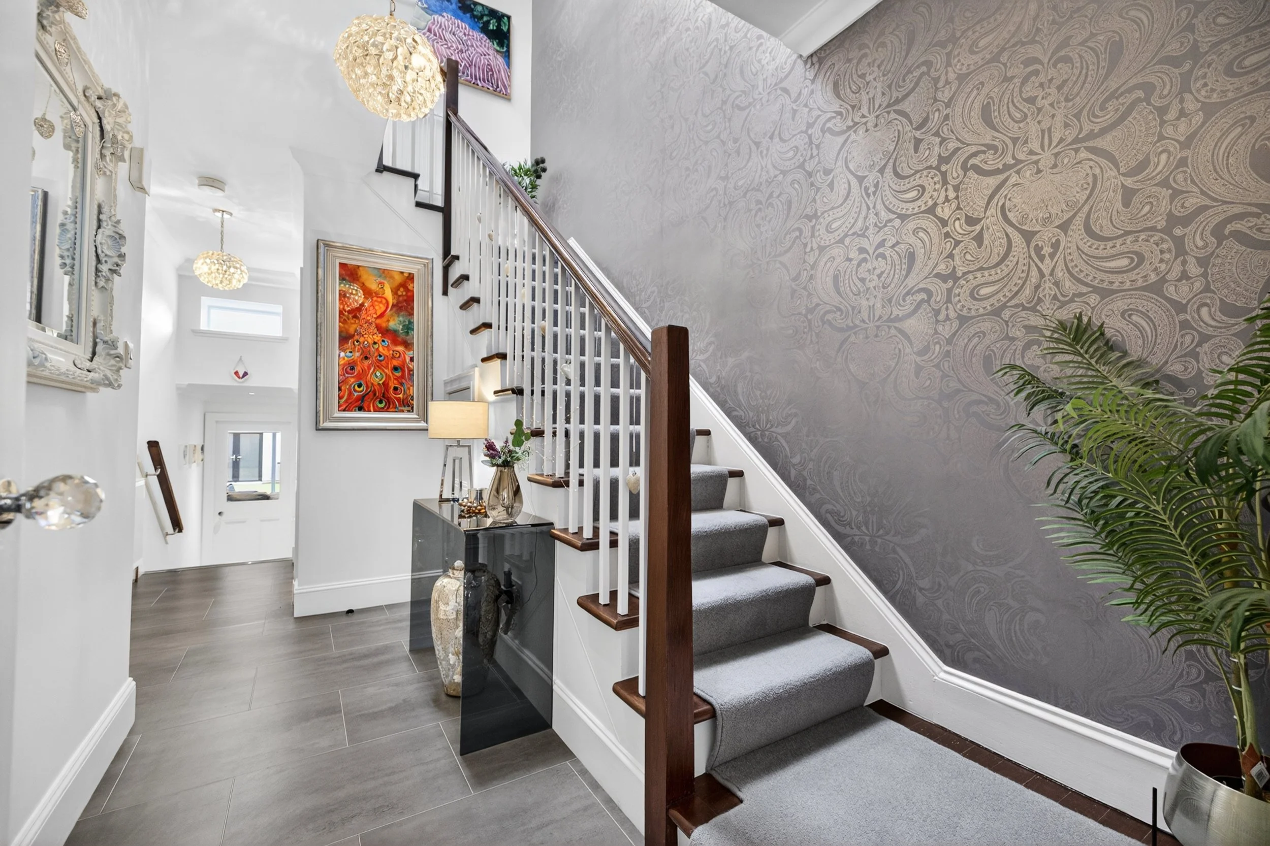 Interior view of a modern staircase area with gray carpeted stairs, a dark gray console table with decorative items, a large ornate mirror, framed artwork including a peacock painting, white walls with decorative molding, a textured gray wall on the 
