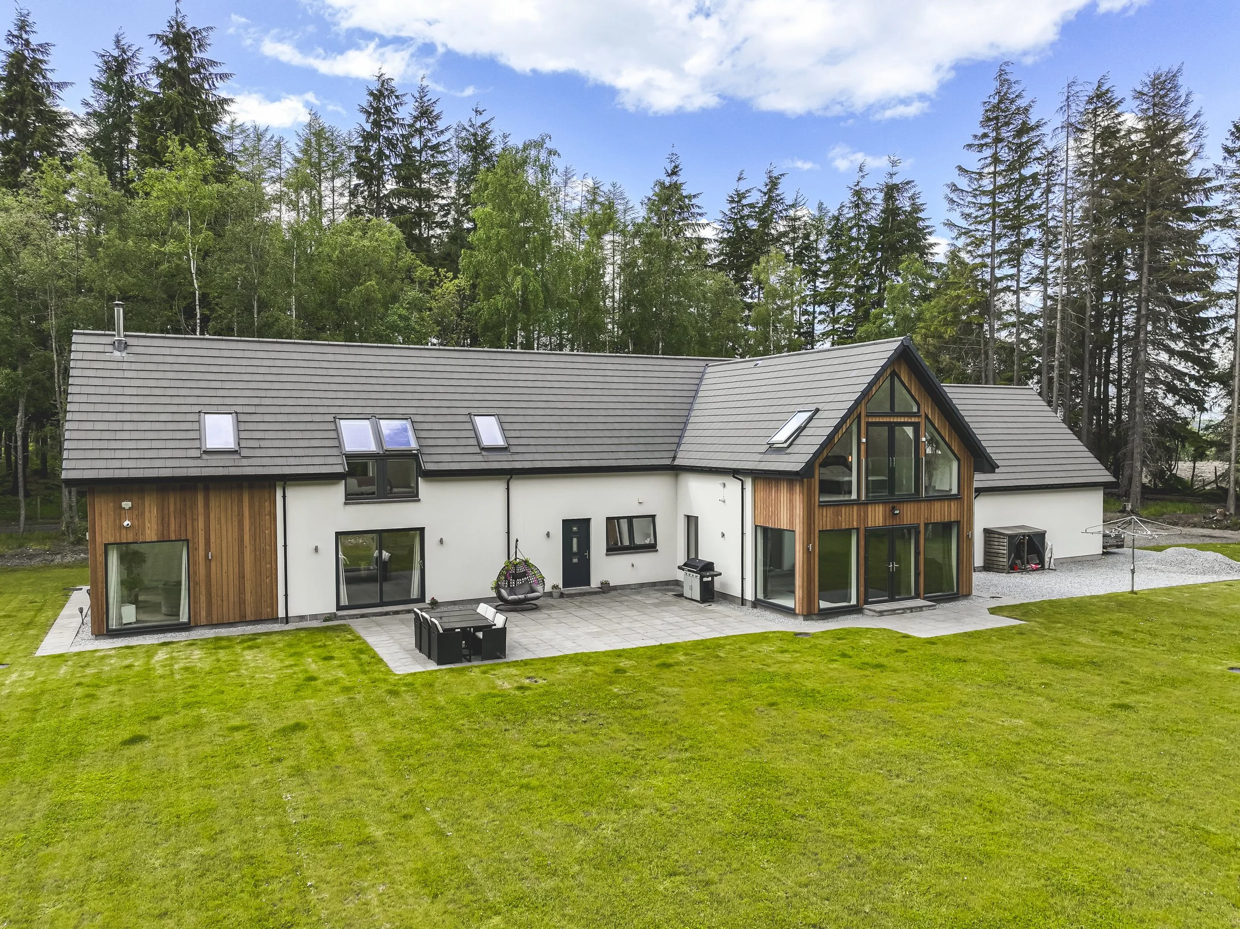 Modern house with a gray sloped roof, large windows, wooden and white exterior walls, surrounded by lush green lawn and tall pine trees in the background under a partly cloudy sky.