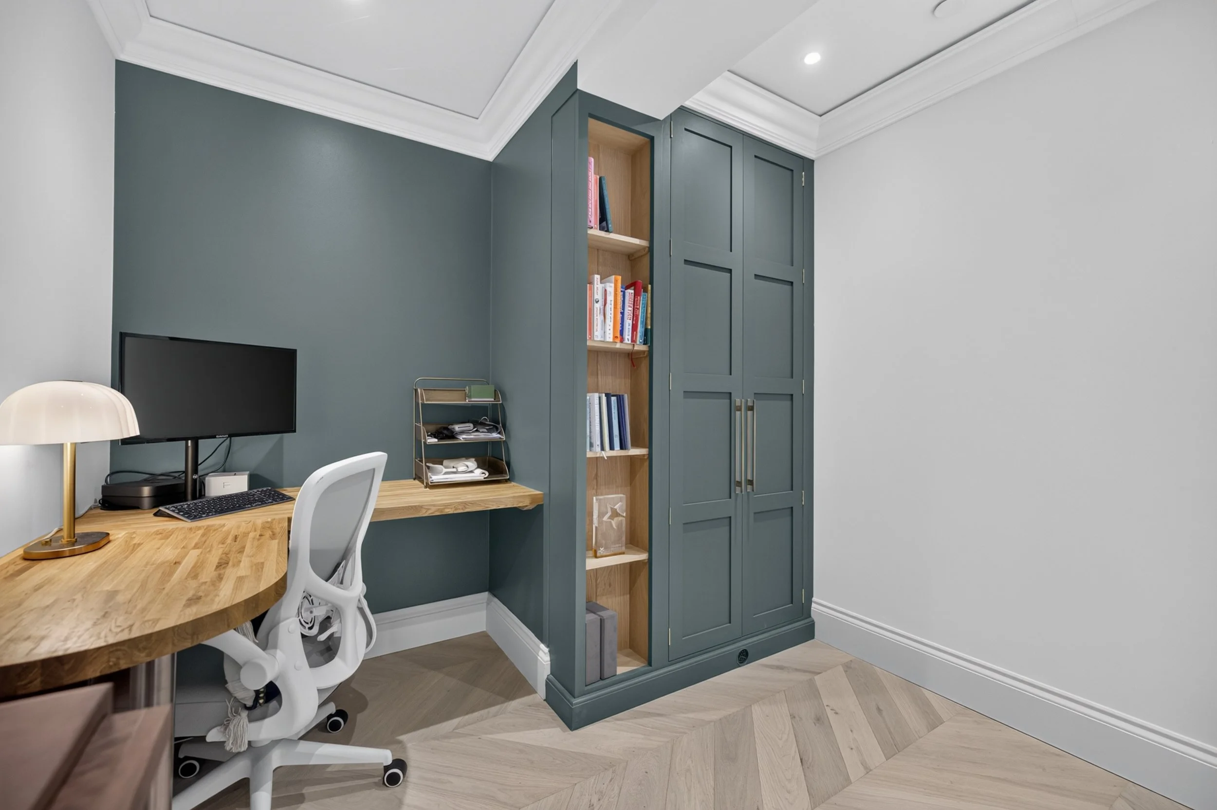 Home office with desk, computer monitor, white ergonomic chair, green accent wall, blue built-in bookshelf, and white ceiling with crown molding.