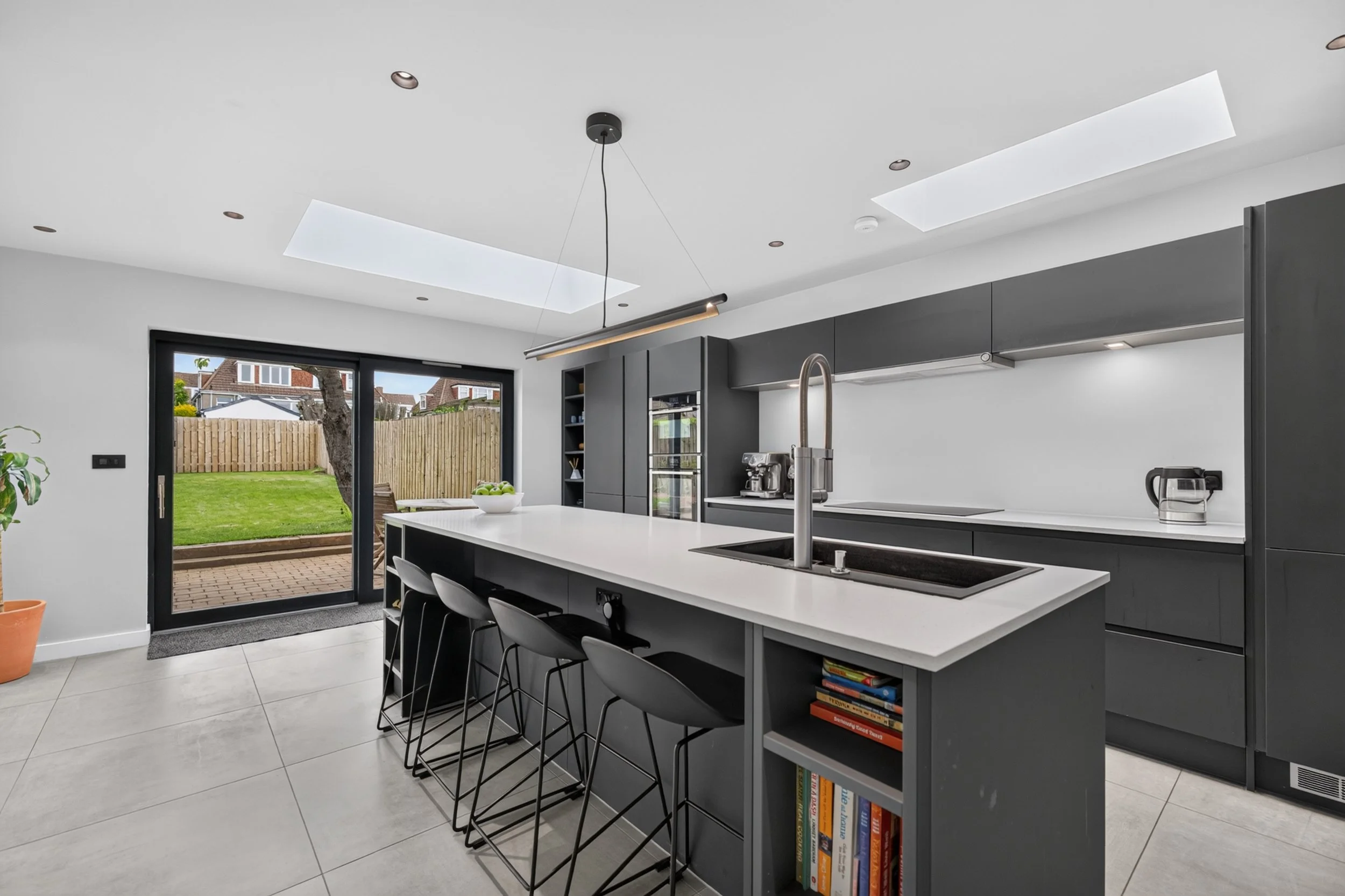 Modern kitchen with white island, dark gray cabinets, sliding glass door to backyard, skylights, and bar stools.