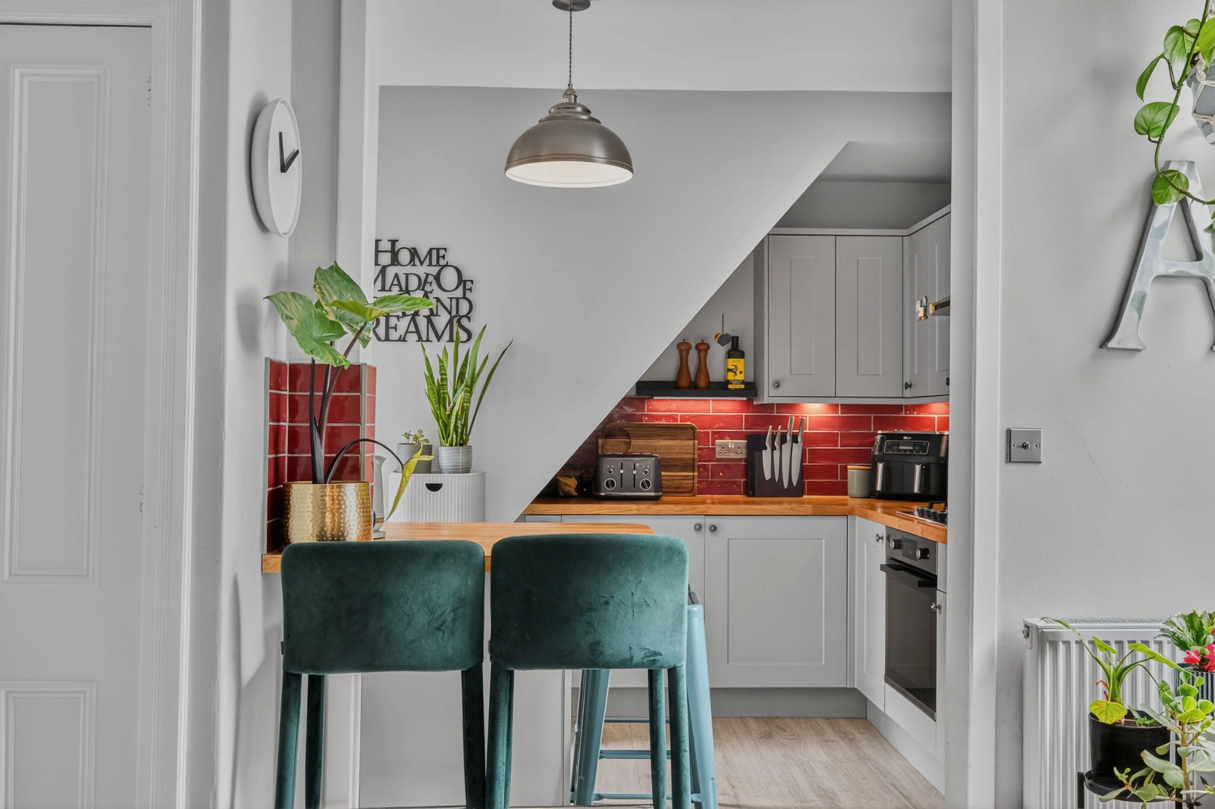 A kitchen with gray cabinets, a red backsplash, and a wooden countertop. There are plants on the counter and a wall sign that reads 'Home Made of Land Dreams.' A ceiling light hangs above the center of the space, and a clock on the wall shows approxi