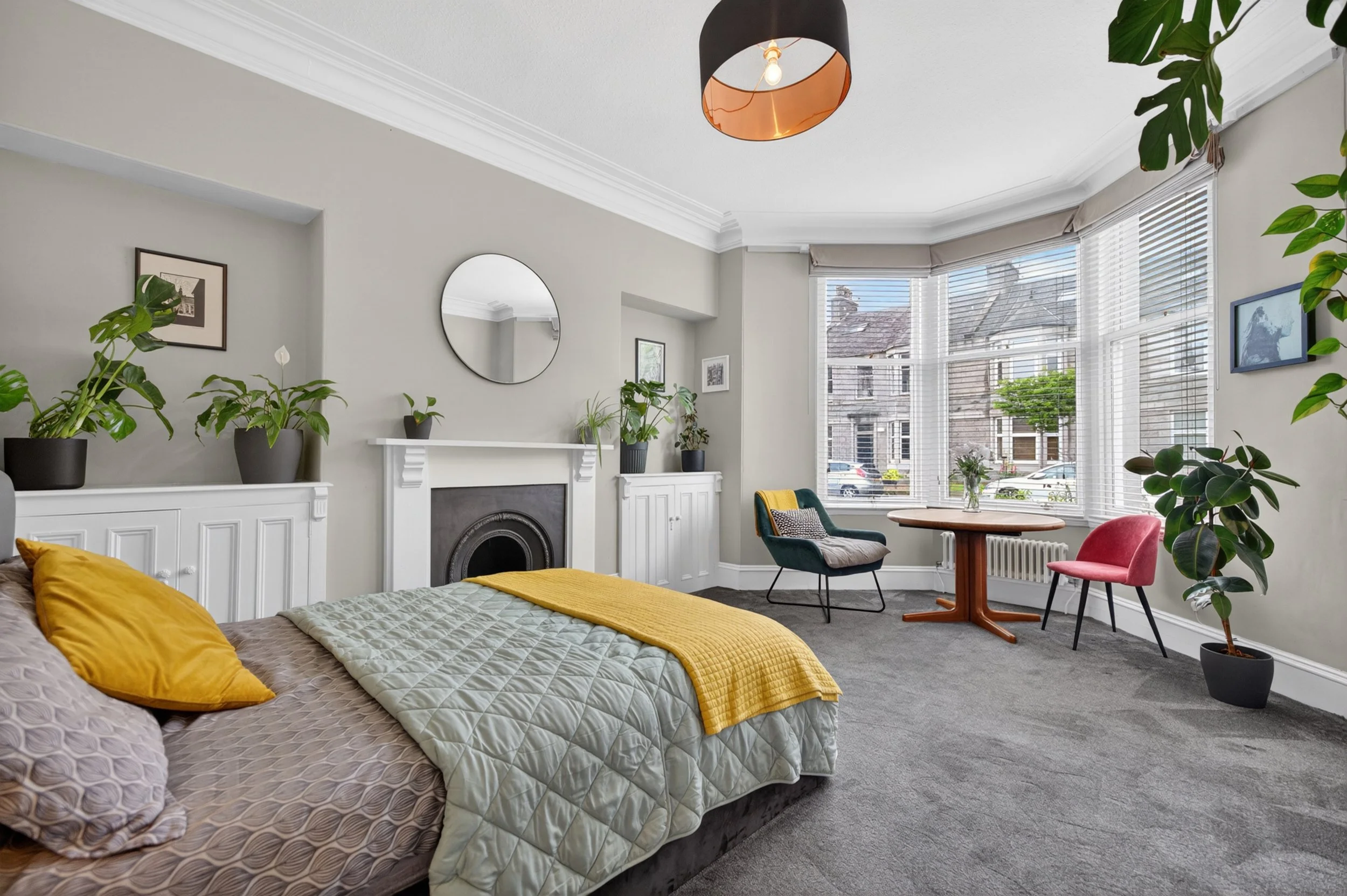 A living room with a large window, plants, a fireplace, a round table, and colorful chairs.