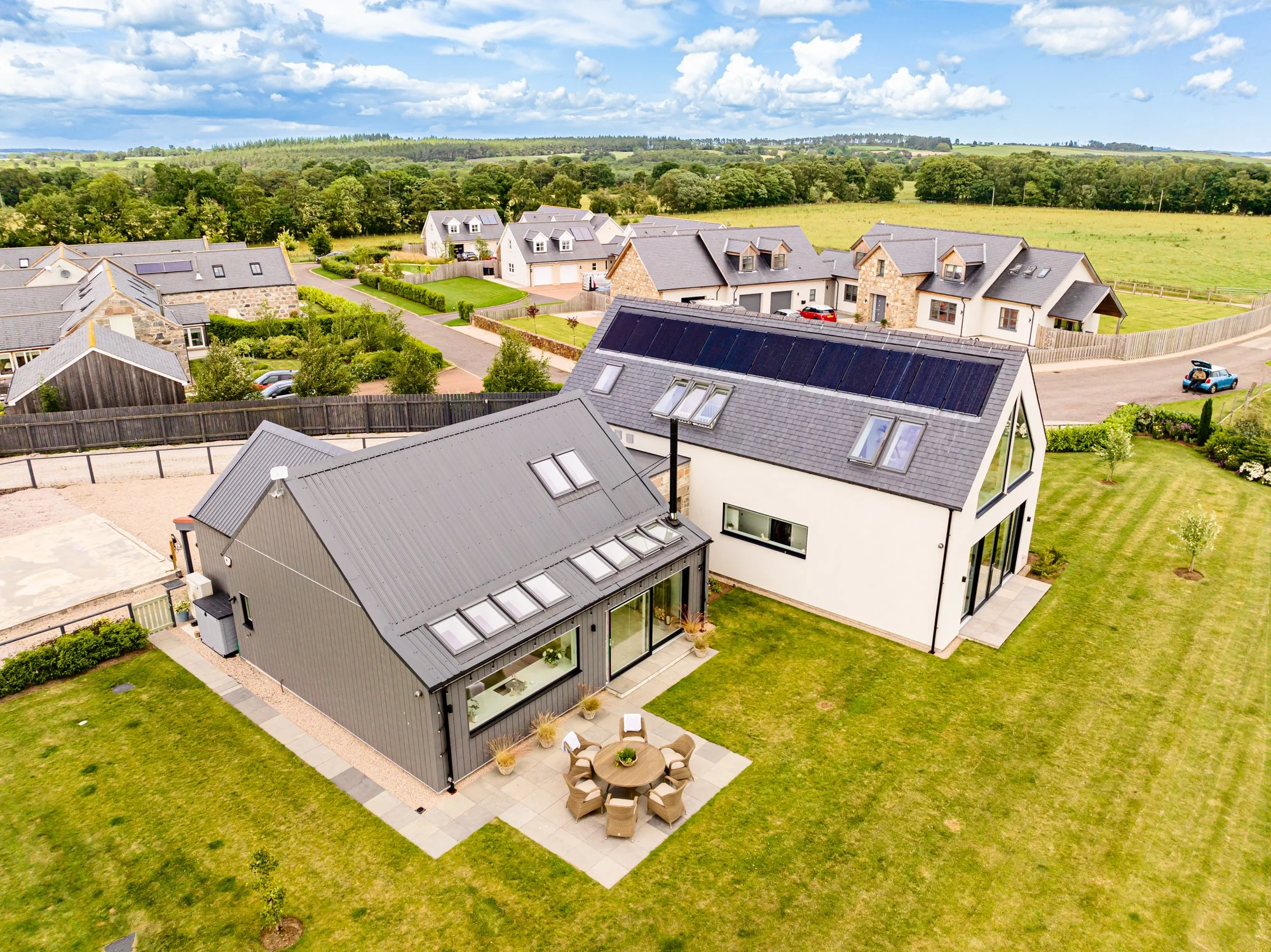 Aerial view of a modern residential neighborhood with several houses featuring gray roofs and solar panels, surrounded by green lawns, trees, and open fields under a partly cloudy sky.