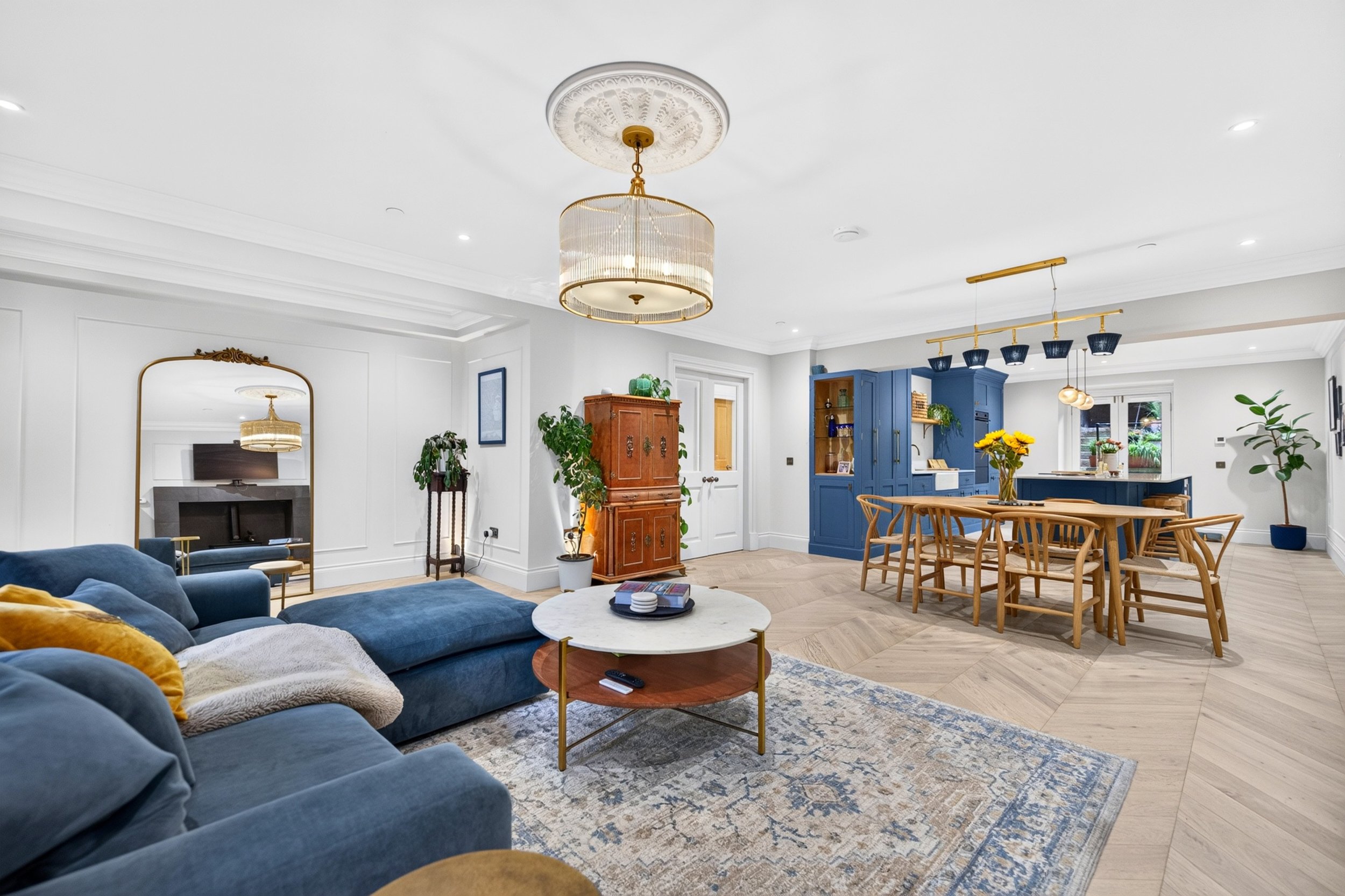 A spacious living and dining area with white walls, light-colored hardwood floors, a blue sectional sofa, a round marble coffee table, a wooden dining table with matching chairs, a blue kitchen cabinet, a gold chandelier, ceiling lights, and various decorative plants.