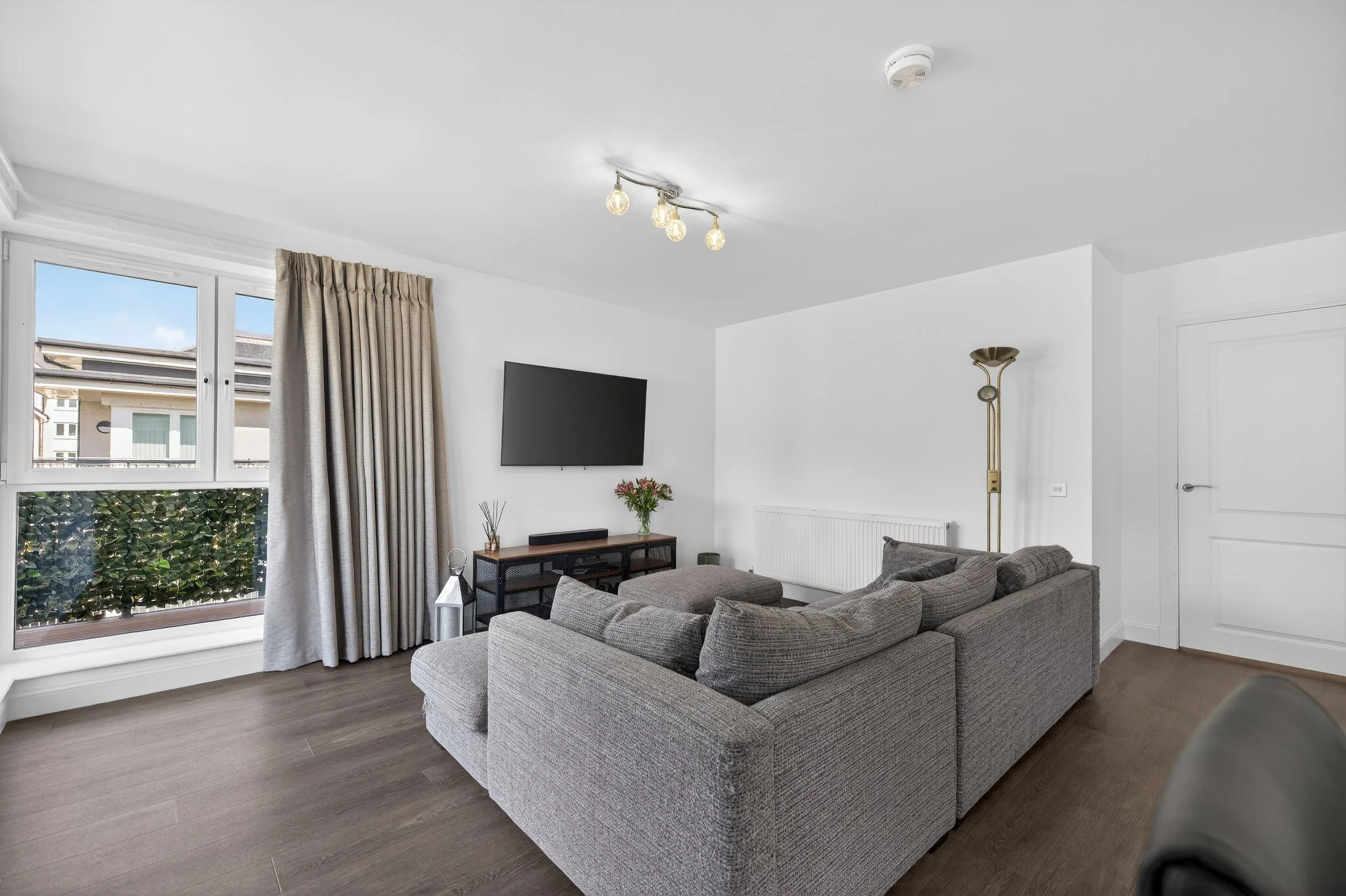 Living room with large windows, gray sectional sofa, flat-screen TV on the wall, beige curtains, wooden floor, and minimalist decor.