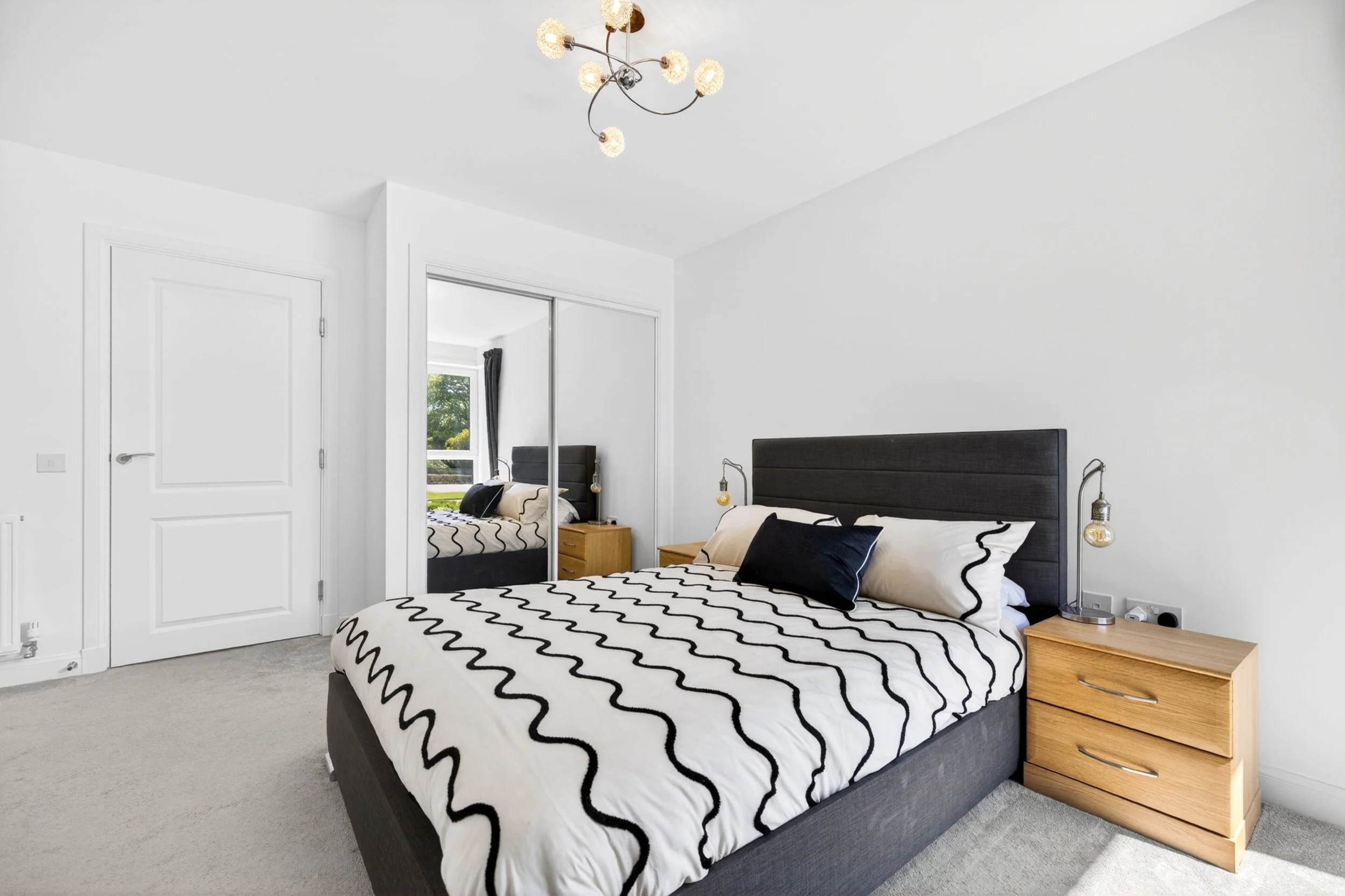 A bedroom with a black bed frame, black and white zigzag patterned bedding, a wooden nightstand, hanging light bulbs on each side of the bed, and a large window with dark curtains.