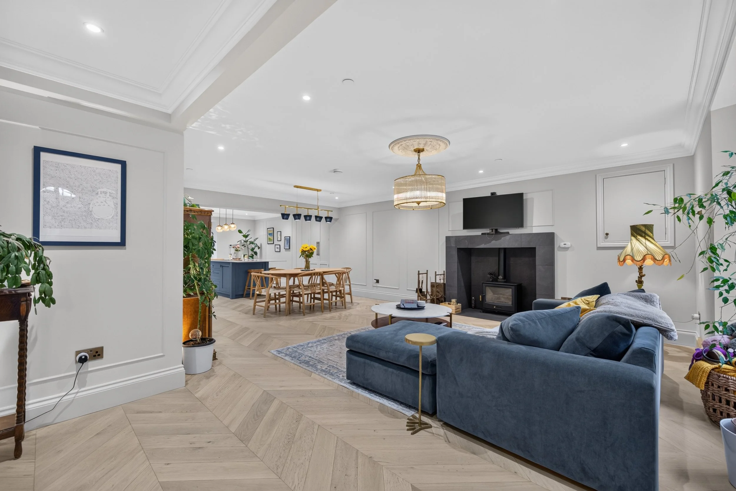 A spacious, modern living room with white walls, light wood flooring, a blue velvet sofa, a round coffee table, potted plants, a flat-screen TV above a fireplace, and a chandelier hanging from the ceiling.