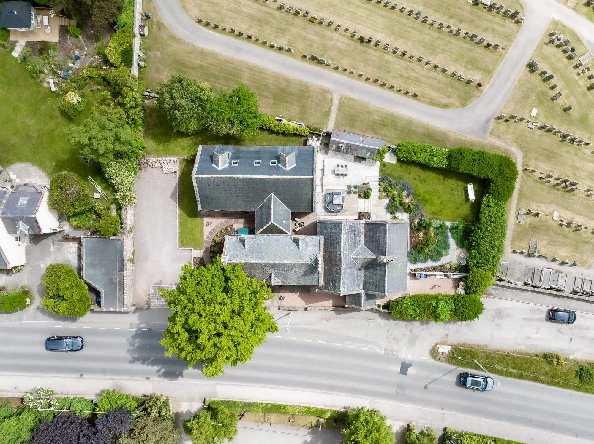 An aerial view of a residential neighborhood showing a house with a dark roof, surrounded by green trees and well-kept lawns. To the right, there is a parking lot with numerous cars parked in rows. A curved road runs through the neighborhood, with a 