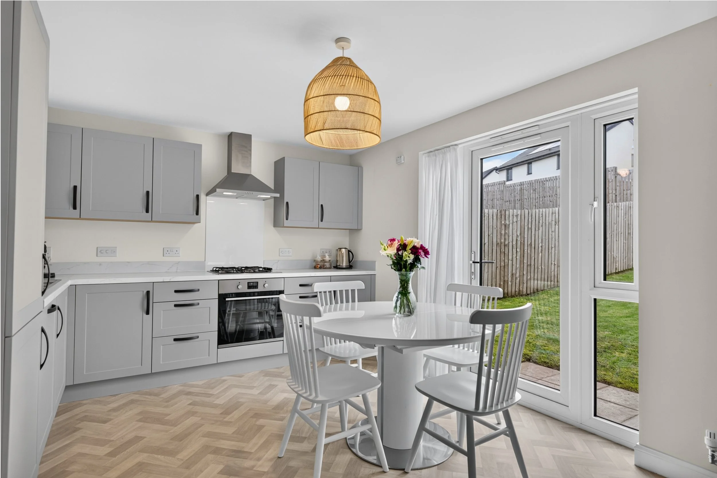 Modern kitchen with white cabinetry, a white round dining table with four chairs, a vase of flowers, and large glass doors leading to a backyard with a wooden fence and grass. A woven pendant light hangs from the ceiling.