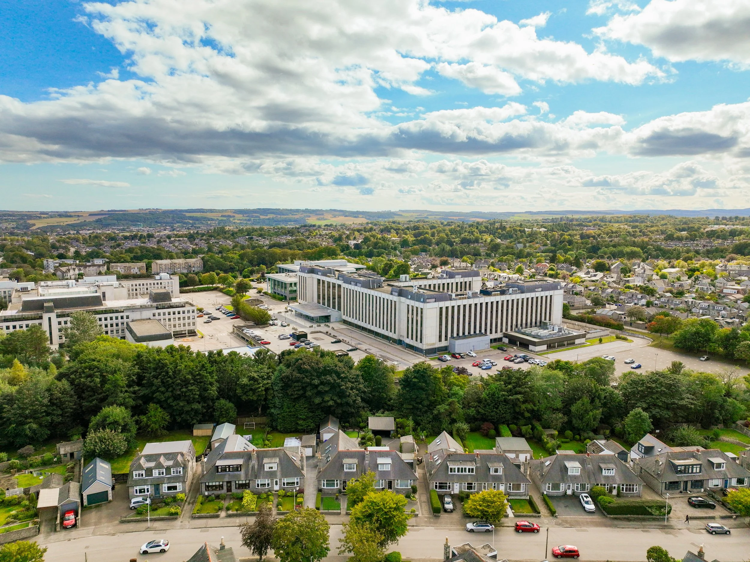 Aerial view of a cityscape with a large modern office building in the foreground, surrounded by residential houses with green lawns, and a vast landscape of trees, fields, and cloudy blue sky in the background.