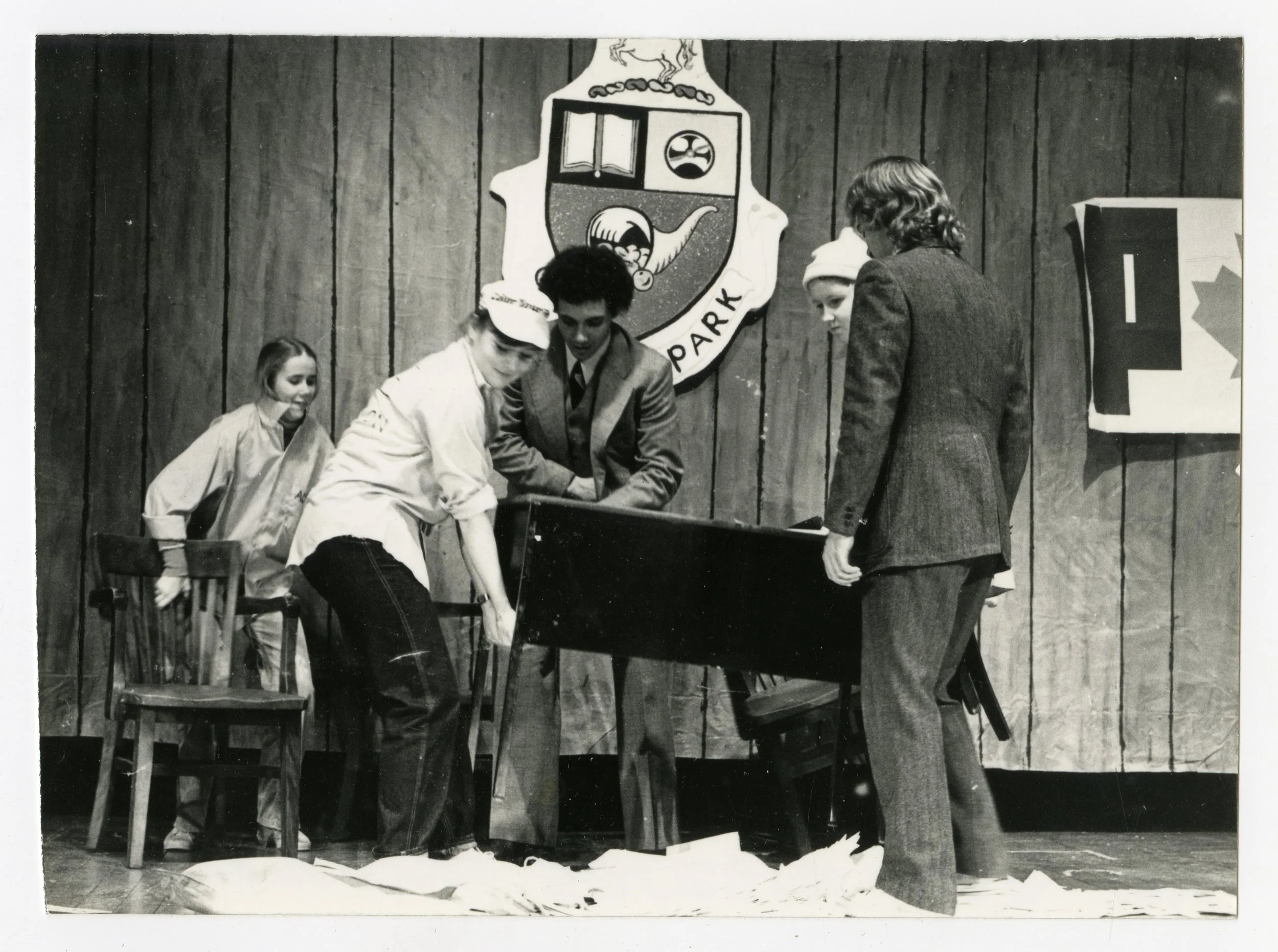 Black and white photo of a group of young people performing a scene on stage. They are gathered around a table, with majority of them picking it up to carry it out. Behind them is the crest of Queen's Park.