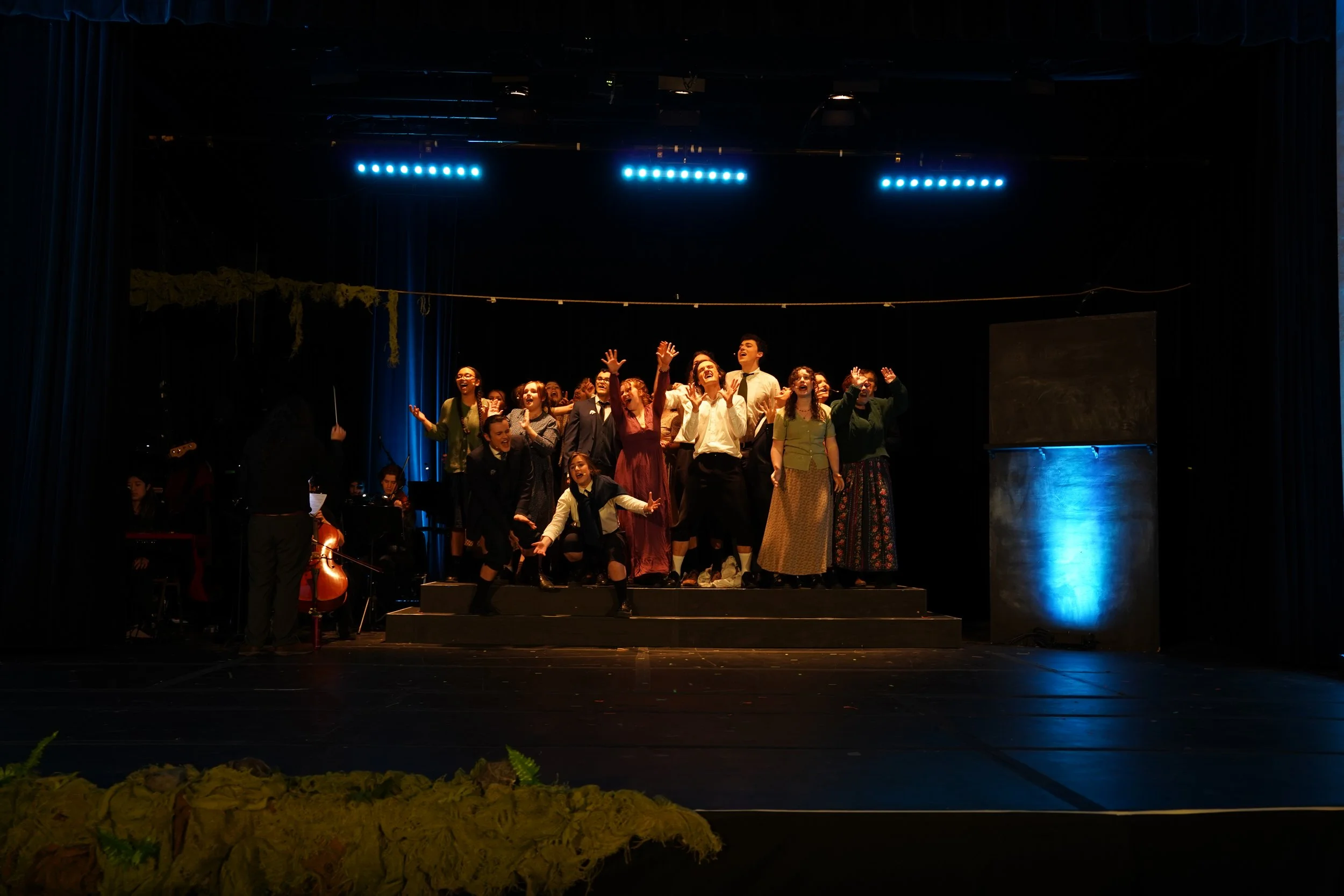 Group of people performing on stage, some singing and dancing, with an orchestra in the background, stage lighting with blue hues, in a theatrical setting.