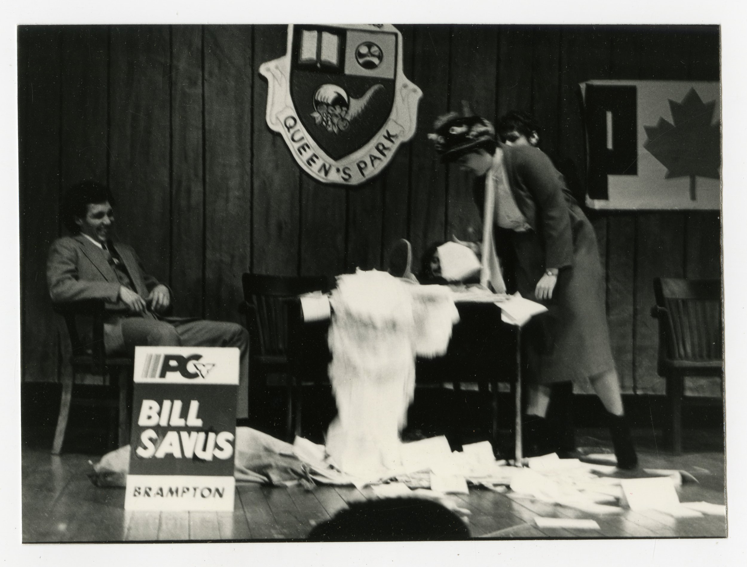 A scene from a political office, with a woman wearing a mask looking at papers on a desk surrounded by scattered documents, while two men sit and stand nearby. There are signs and posters indicating political campaign signs for Bill Savus from Brampt
