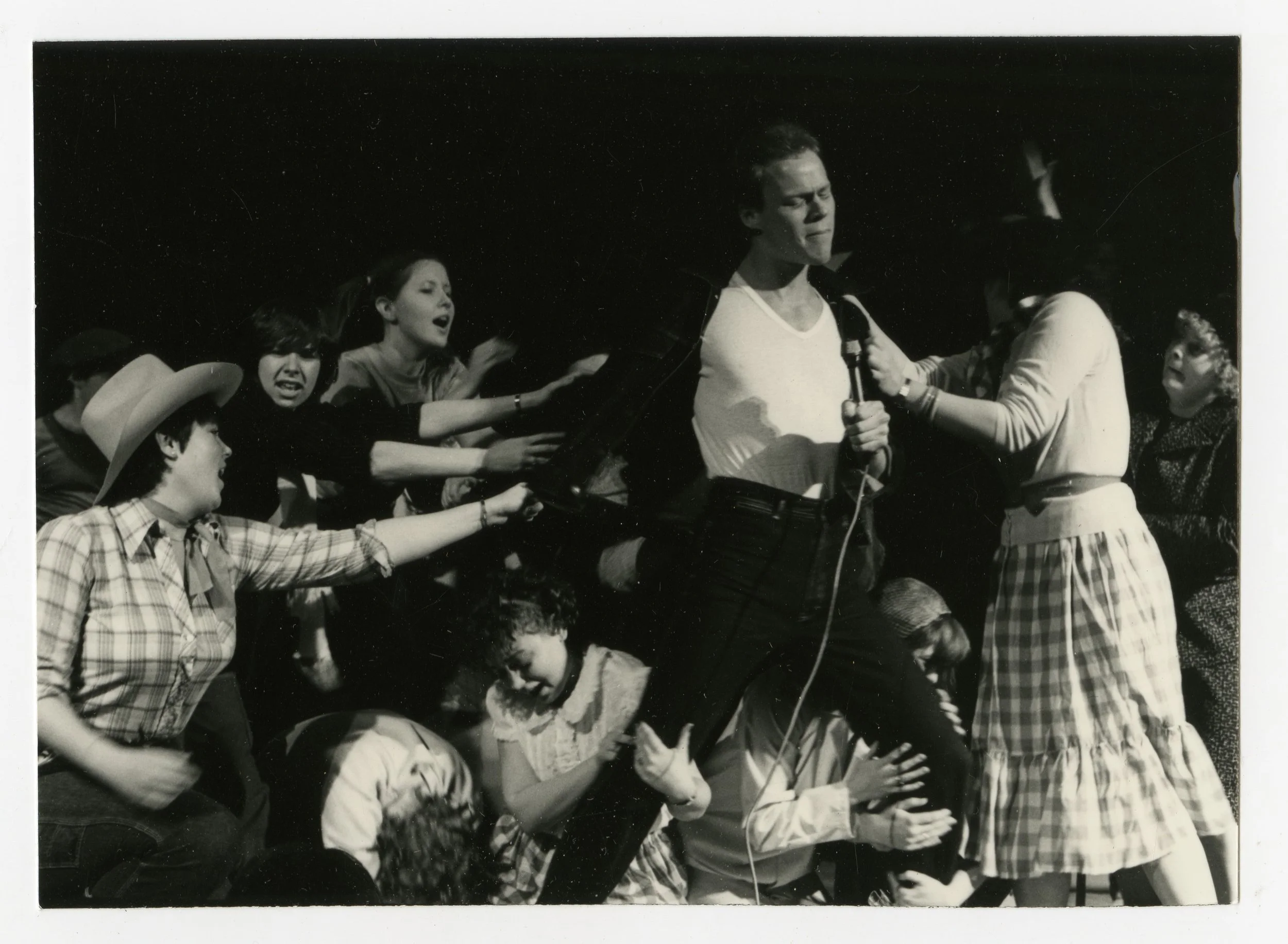 A black and white photo of a man singing or speaking into a microphone, surrounded by a group of characters who are reaching out to grab him and showing various emotional reactions.