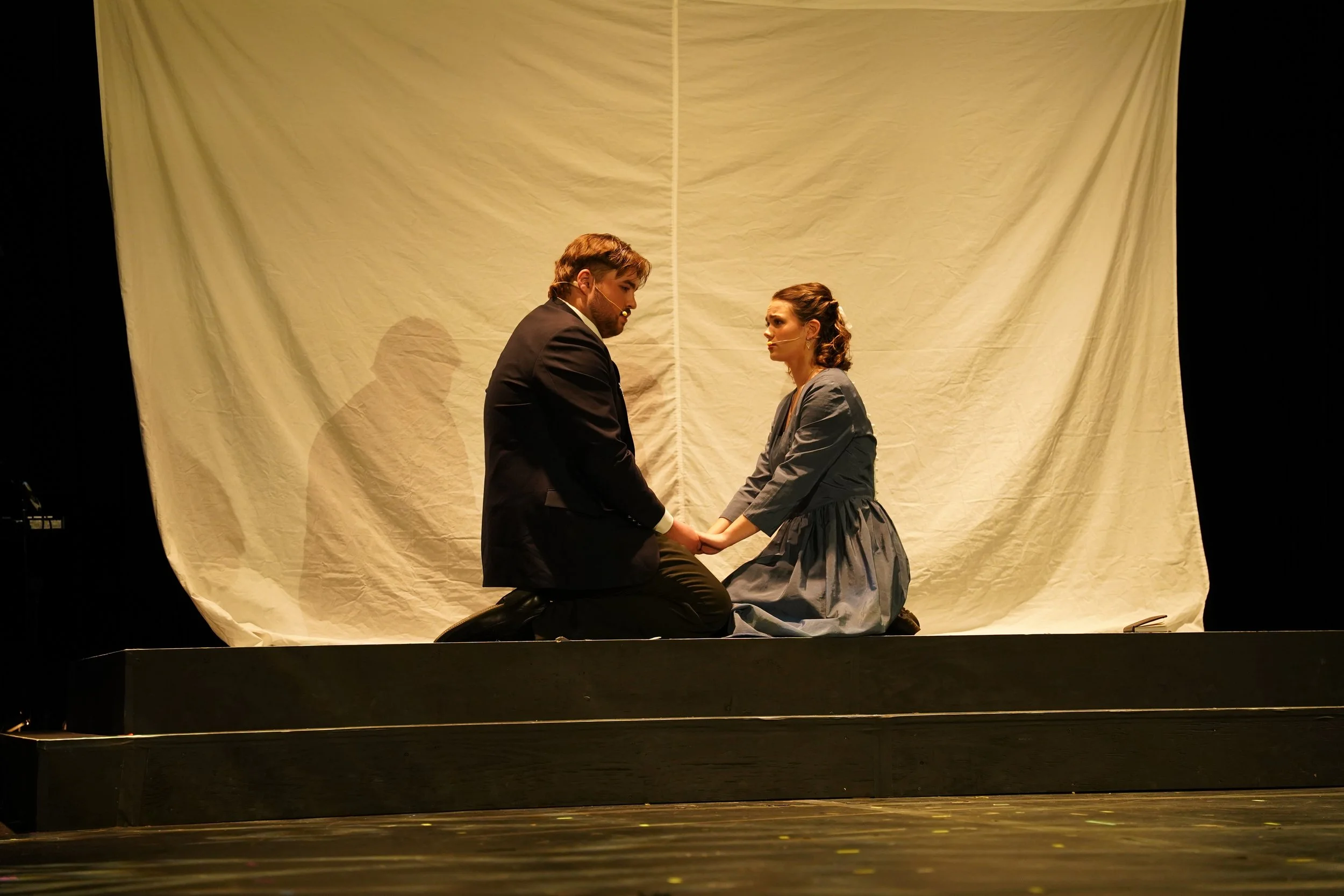 A man and woman kneel on a stage, holding hands and facing each other during a performance, with a white curtain backdrop.