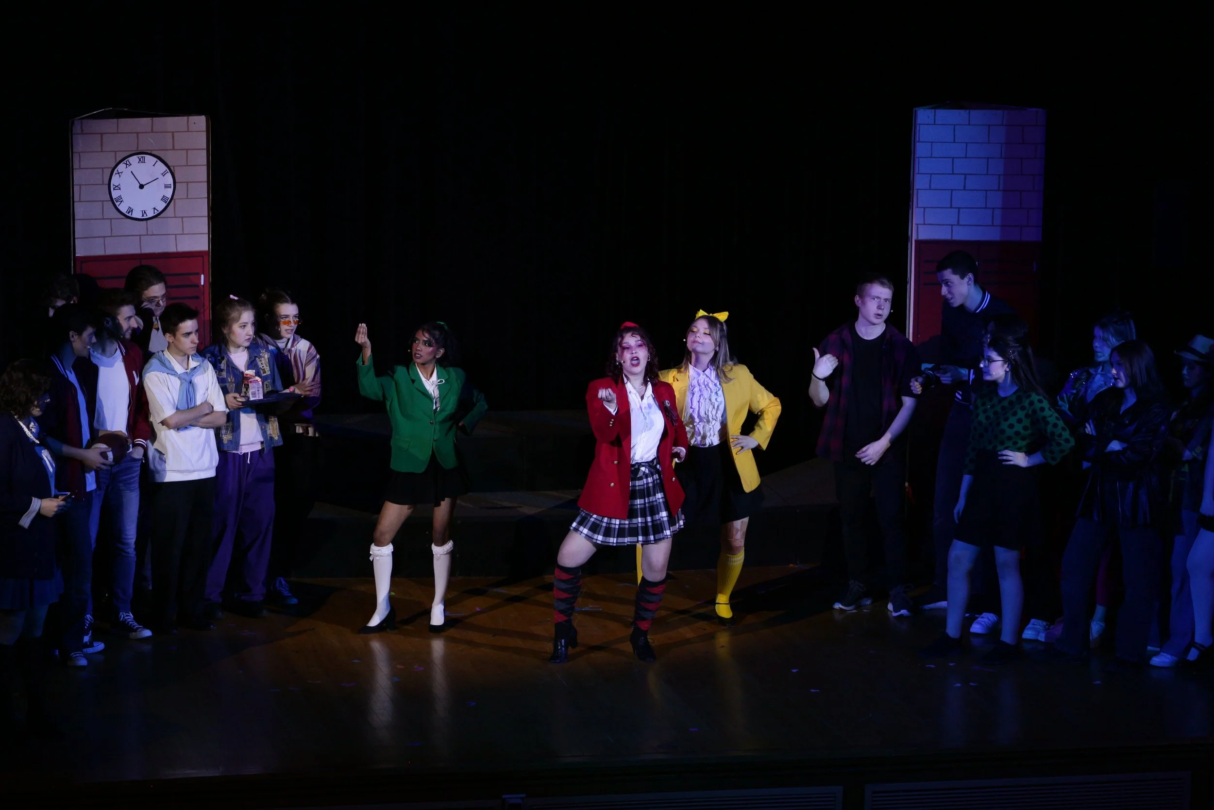 A group of young people performing a play or musical on stage, with some in colorful costumes and others watching.