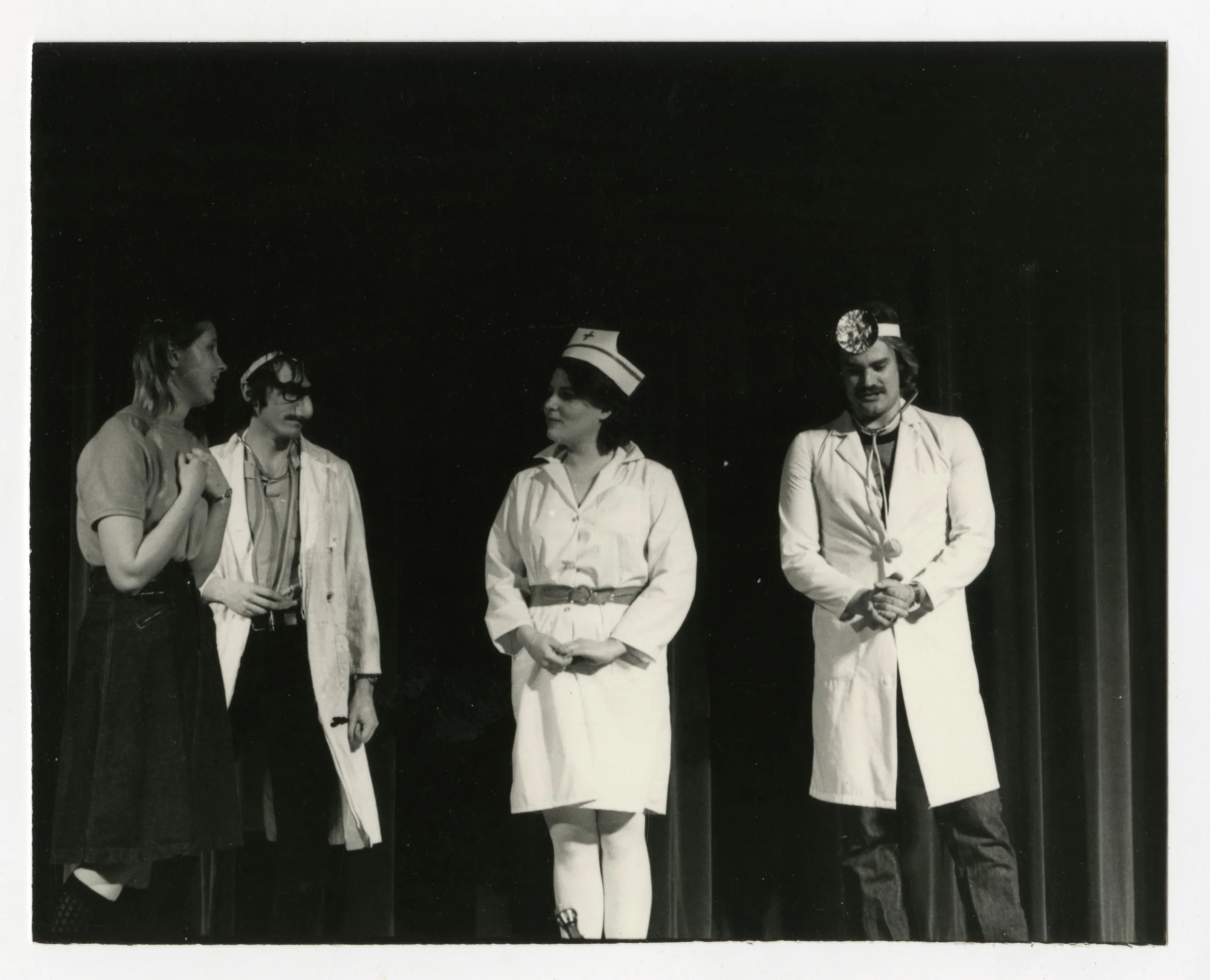 Four people dressed as medical professionals and nurses on stage, part of a performance, with dark curtain in background.