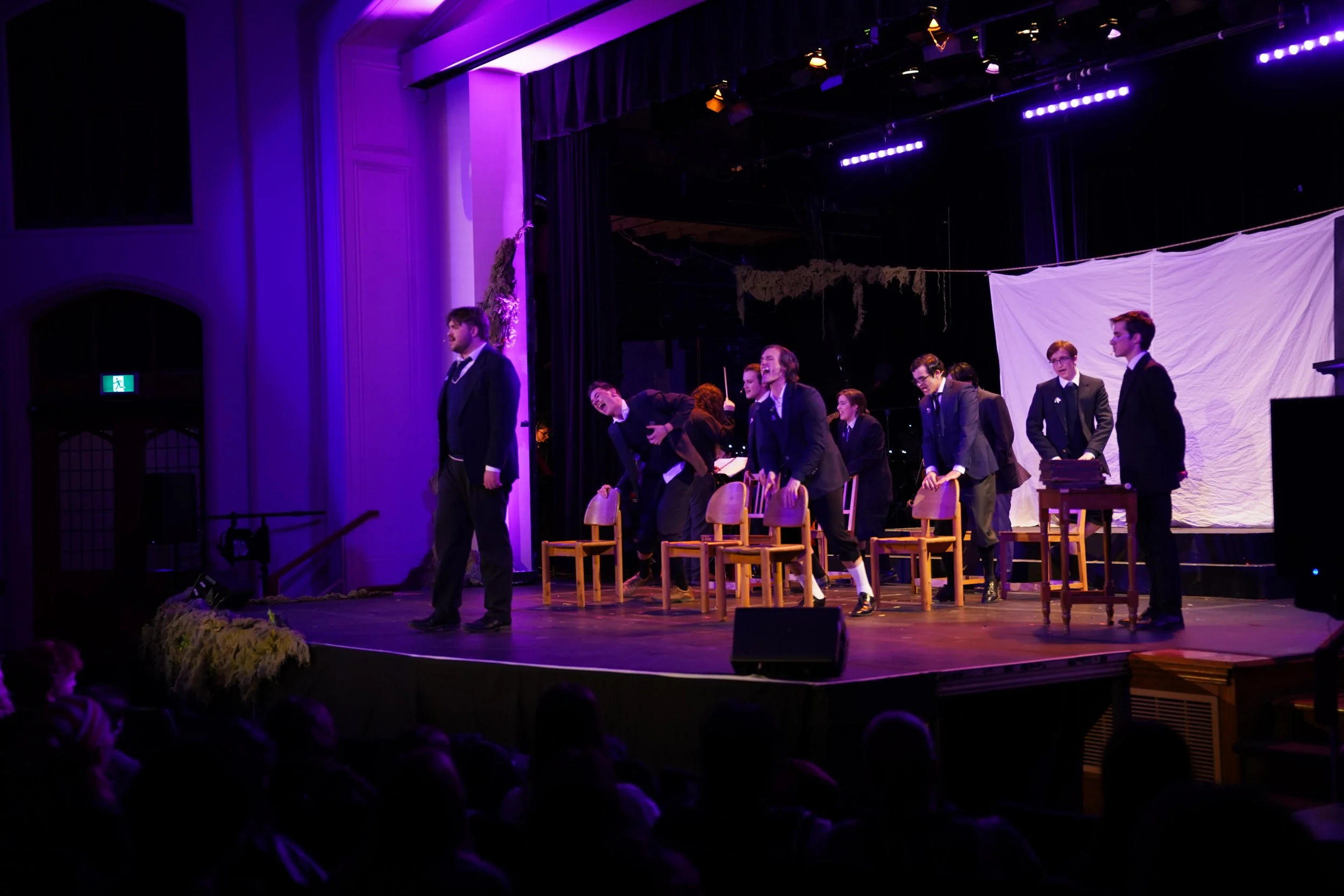 A theatrical performance on stage with actors dressed in formal costumes, some standing and some sitting, with chairs arranged in front of a white backdrop. The stage is lit with purple lighting, and an audience watches from below.