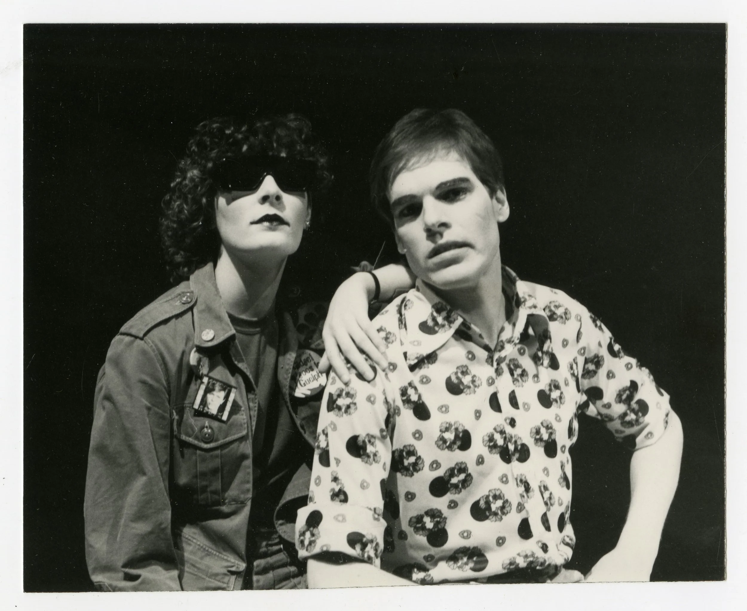Black and white photo of a woman wearing sunglasses and a military-style jacket, and a man wearing a patterned shirt, posing together against a dark background.