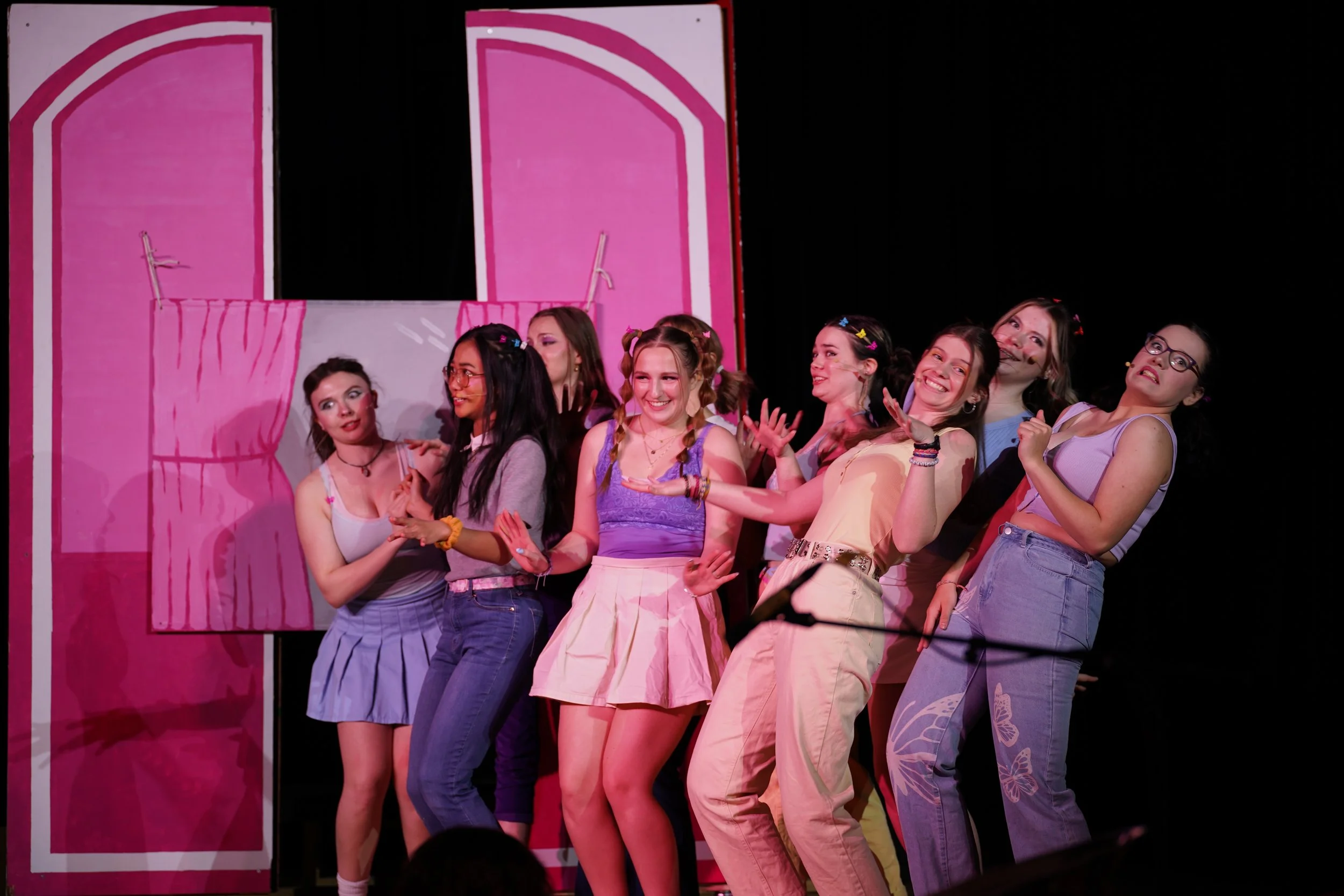 Group of young women performing on stage with a pink set background, some smiling and holding hands.