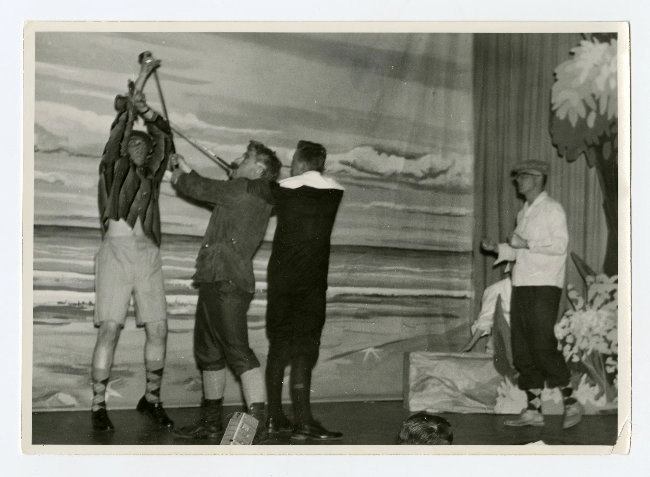 Four men on stage, three holding a flag or banner, with painted scenic backdrop of the ocean and sky, and a man in white coat and cap standing to the right.