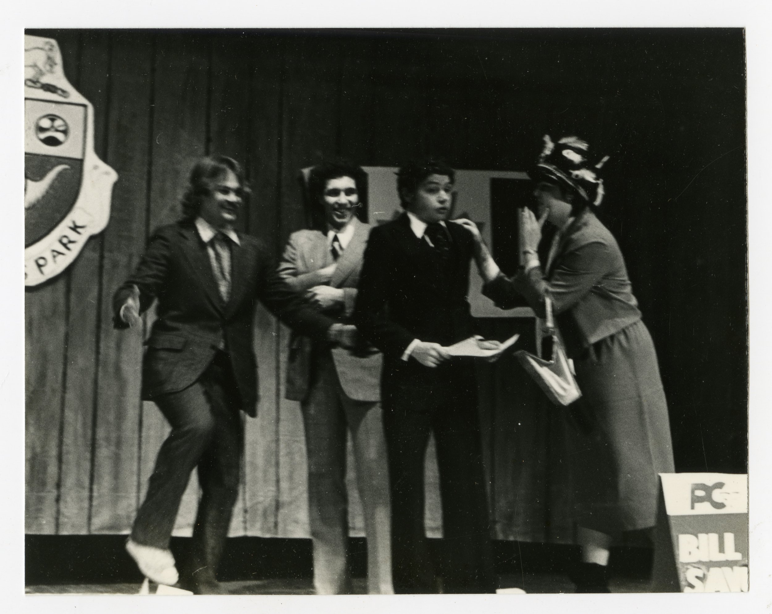 Black-and-white photograph of three boys in suits on stage, talking to a woman wearing a hat and coat, with a wooden background and a crest of Queen's Park on the wall.