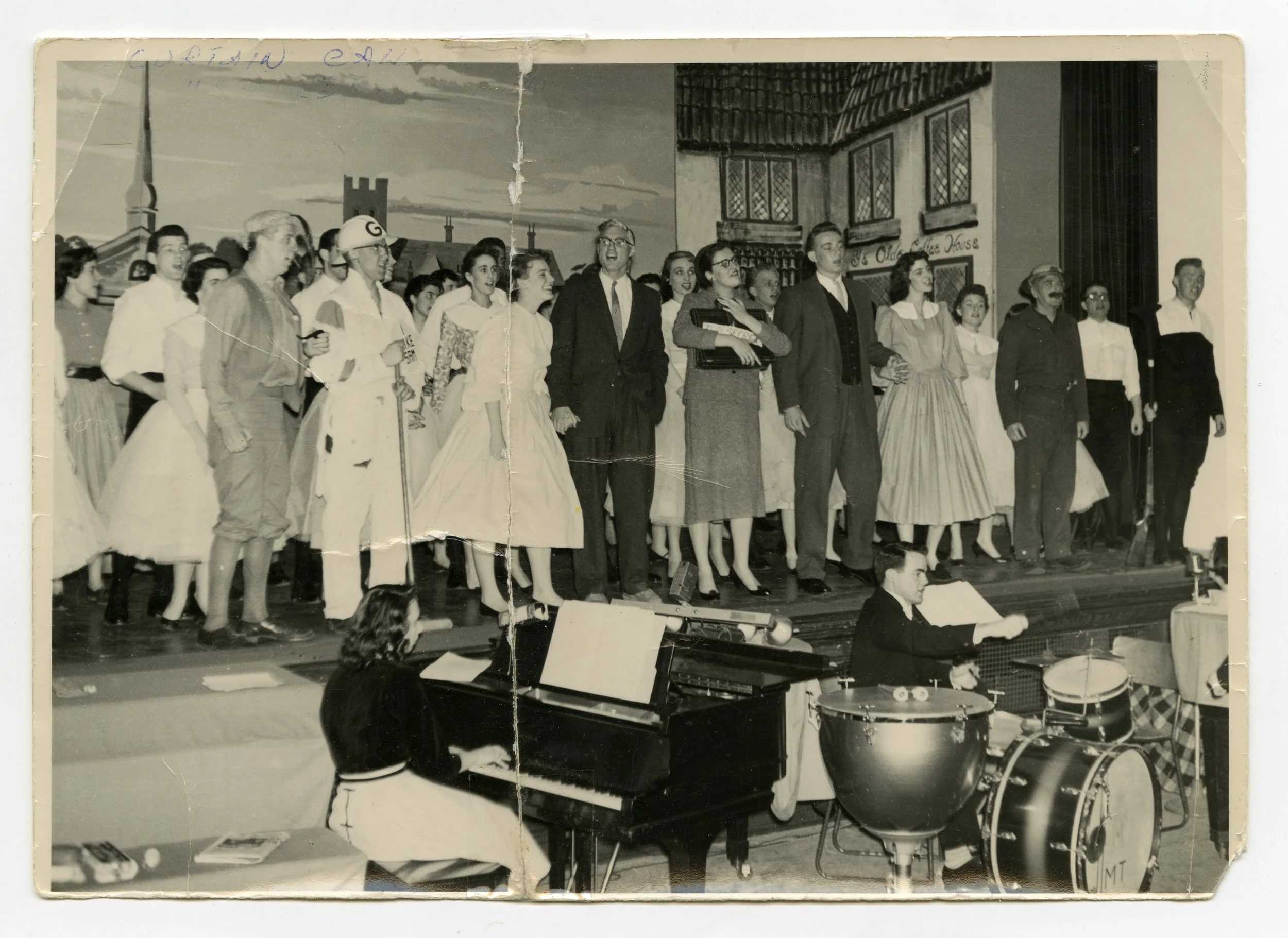 A black and white photo of a theatrical performance with a group of people on stage, including singers and musicians, in a setting resembling a town or village square with painted buildings in the background.