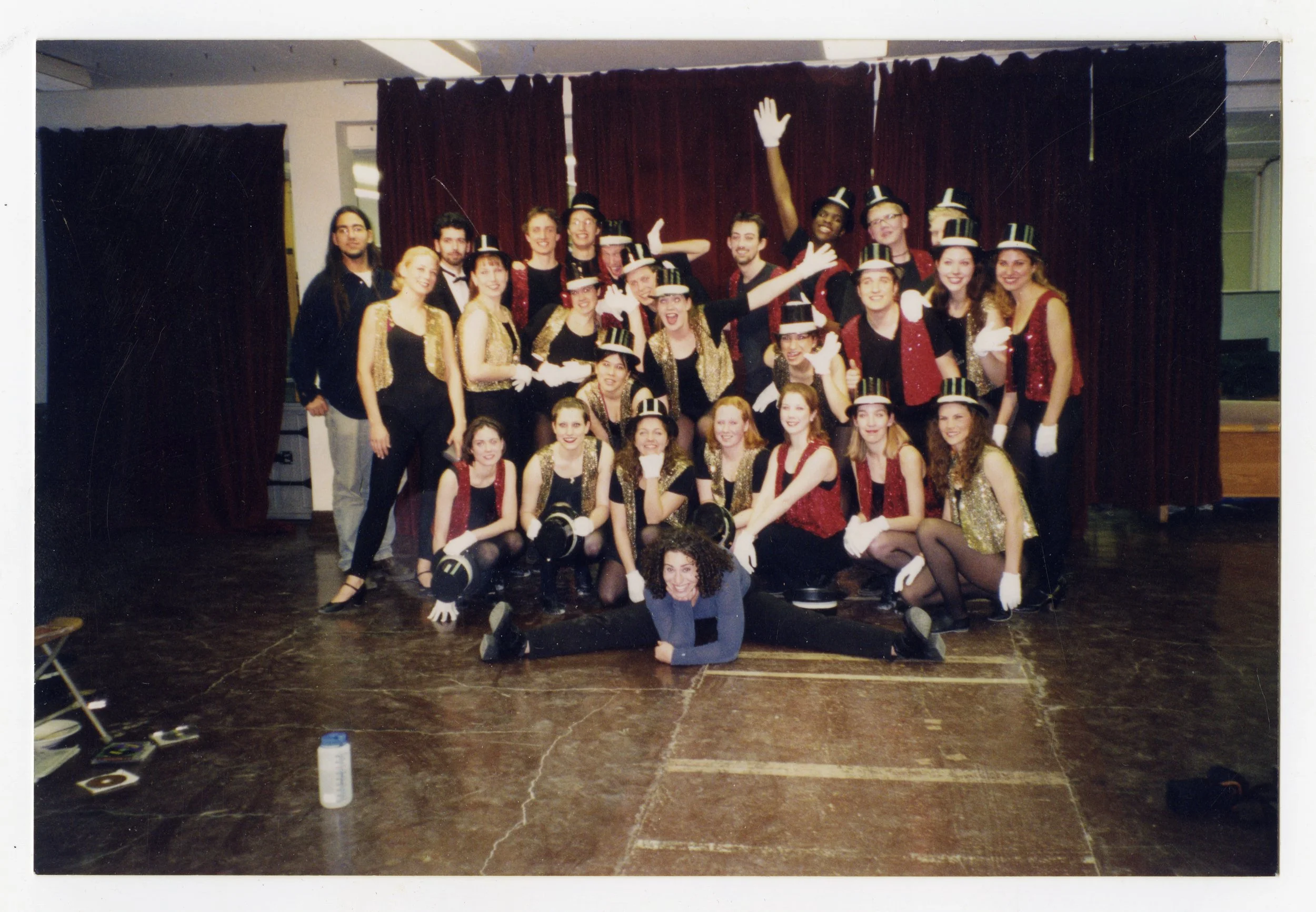 A large group of performers dressed in costumes with black and gold colors, some wearing top hats, gathered on a stage with red curtains behind them. One person is lying on the floor in front, doing a split, and everyone is smiling or making playful 